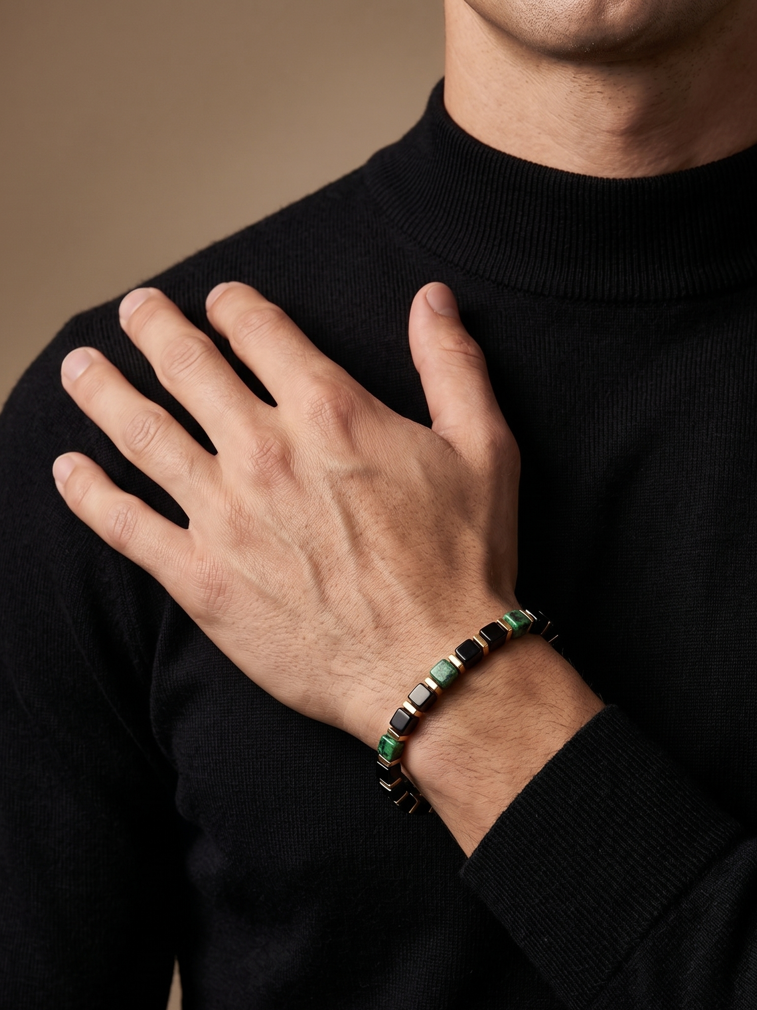 A close-up of a man wearing a black turtleneck sweater and a beaded bracelet on his wrist, with his hand resting on his chest against a warm beige background.