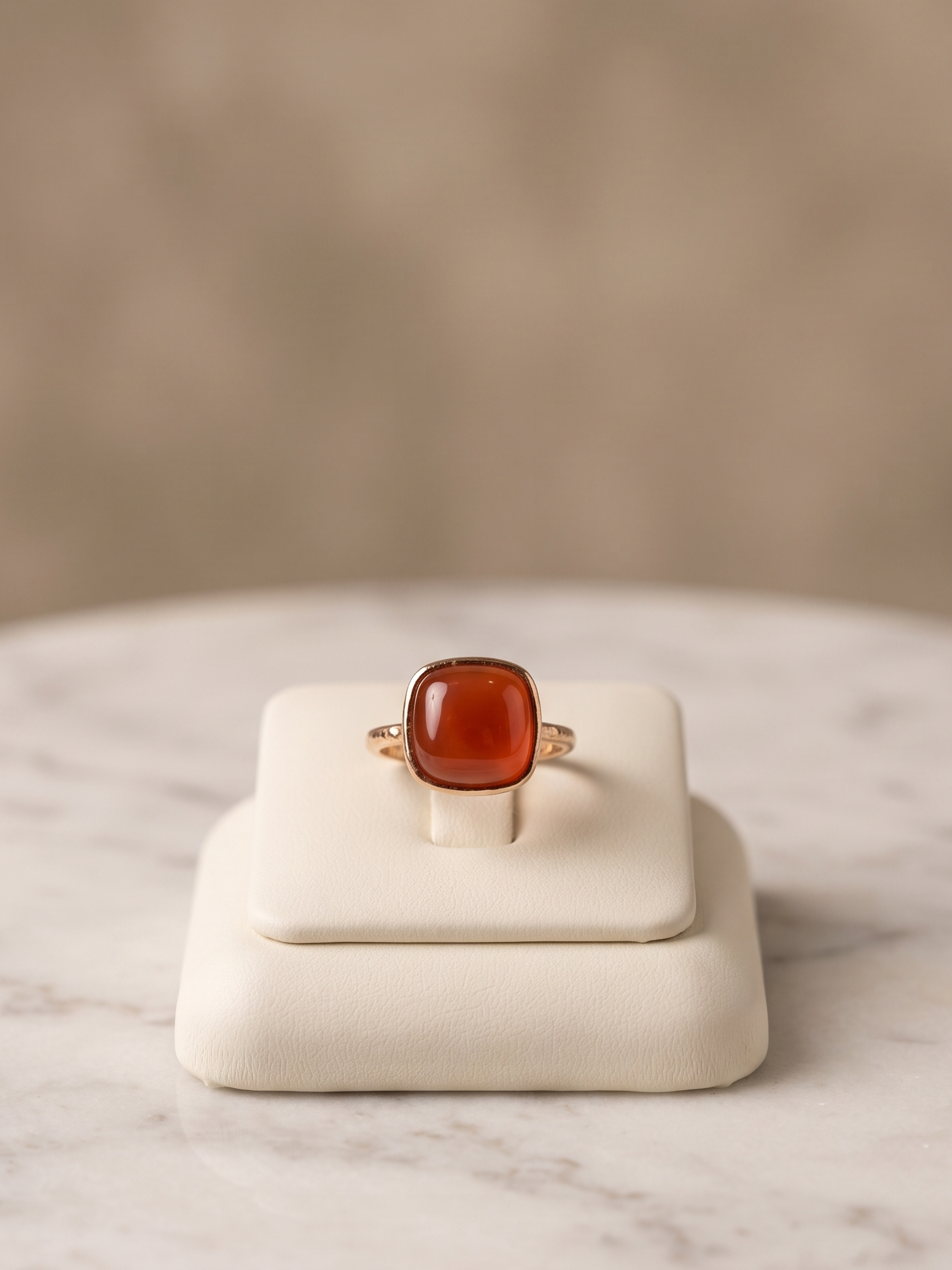 A gold ring with a cushion-cut amber gemstone, displayed on a cream-colored jewelry stand atop a white marble surface with a soft beige background.
