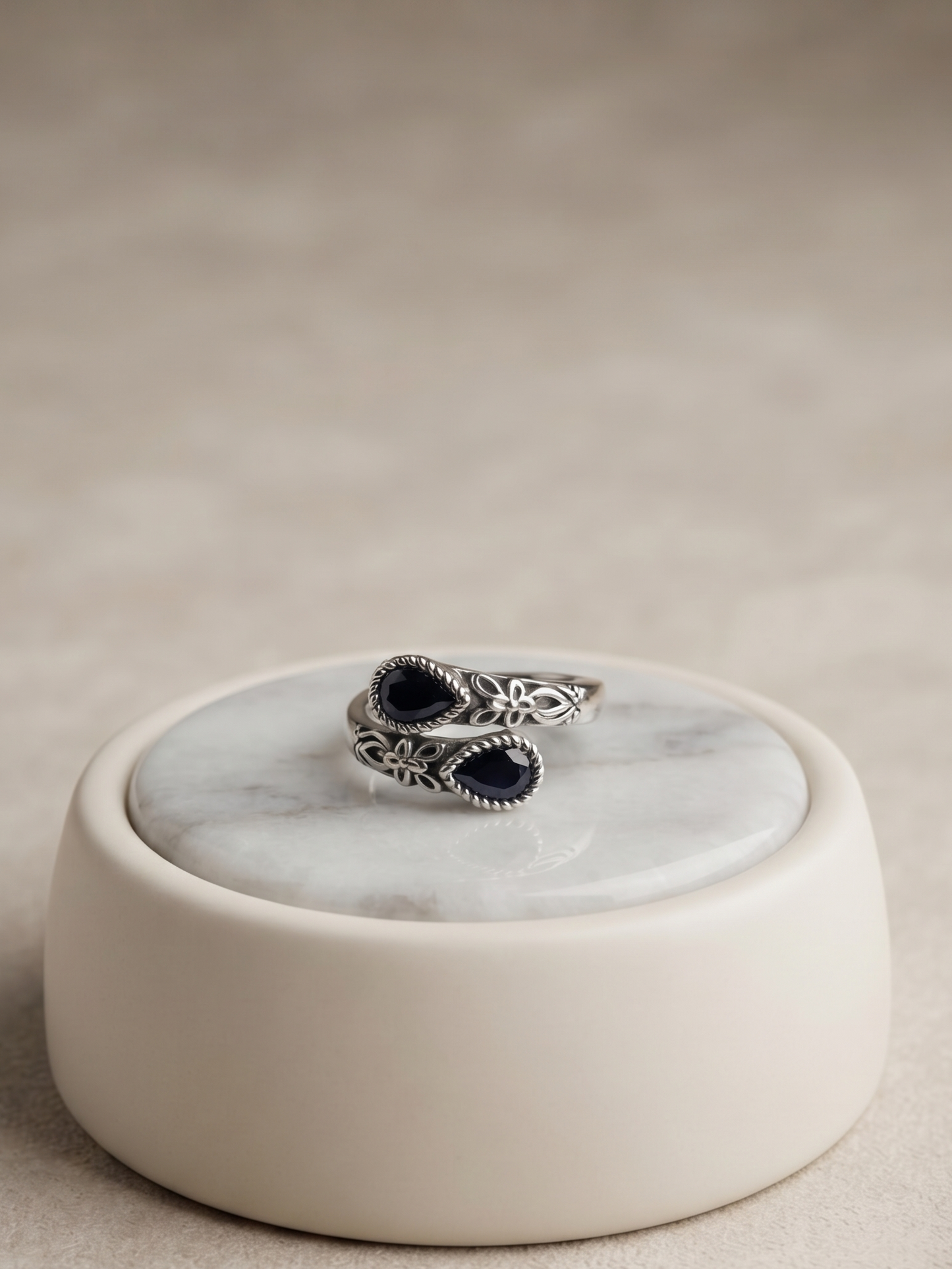 A silver-toned ring with two dark oval stones, displayed on a round cream-colored ceramic stand with a white marble top, set against a neutral beige background.