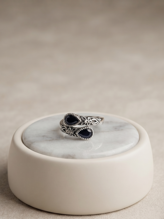 A silver-toned ring with two dark oval stones, displayed on a round cream-colored ceramic stand with a white marble top, set against a neutral beige background.