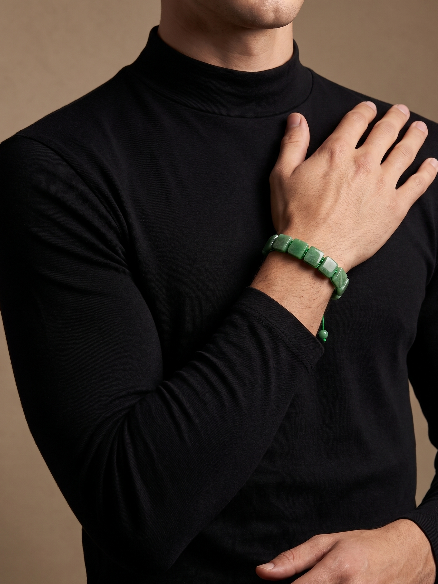 A close-up of a man wearing a black long-sleeve mock neck turtleneck sweater, with his right hand resting on his chest. He is wearing a green beaded bracelet on his wrist, set against a solid warm brown studio background.
