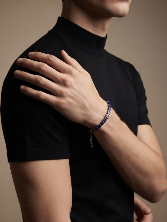 A close-up of a man wearing a black short-sleeve mock neck shirt and a blue beaded bracelet with a silver chain, posed against a solid warm brown background.