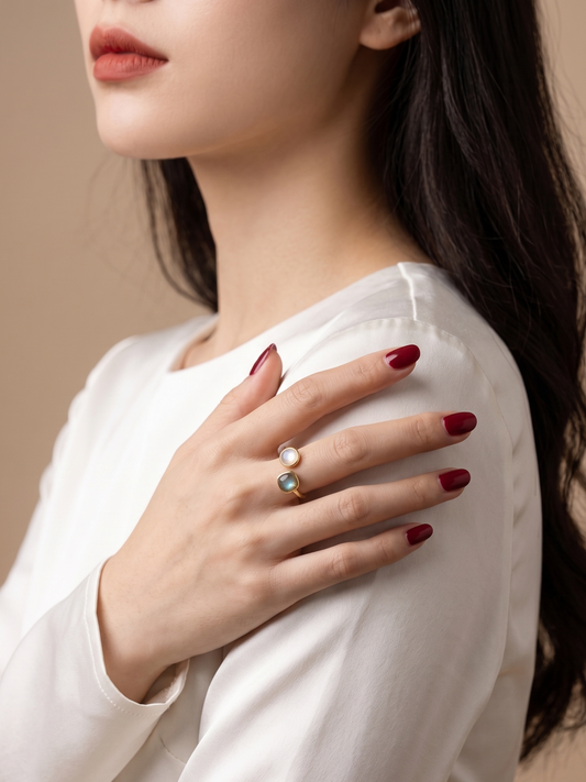 A close-up lifestyle shot of a woman wearing a white long-sleeve top, showcasing two delicate rings on her fingers. The rings feature a small white pearl and a light blue stone on a thin gold band.