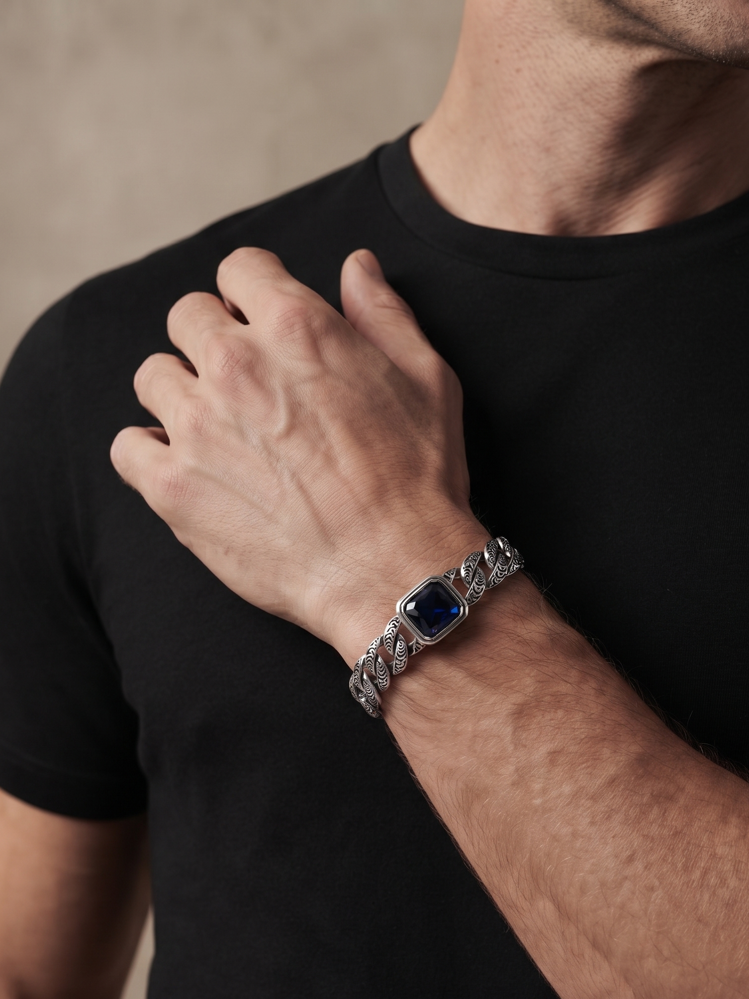 A close-up of a man wearing a silver-toned chain-link bracelet with a square dark blue bezel on his wrist, paired with a black t-shirt.