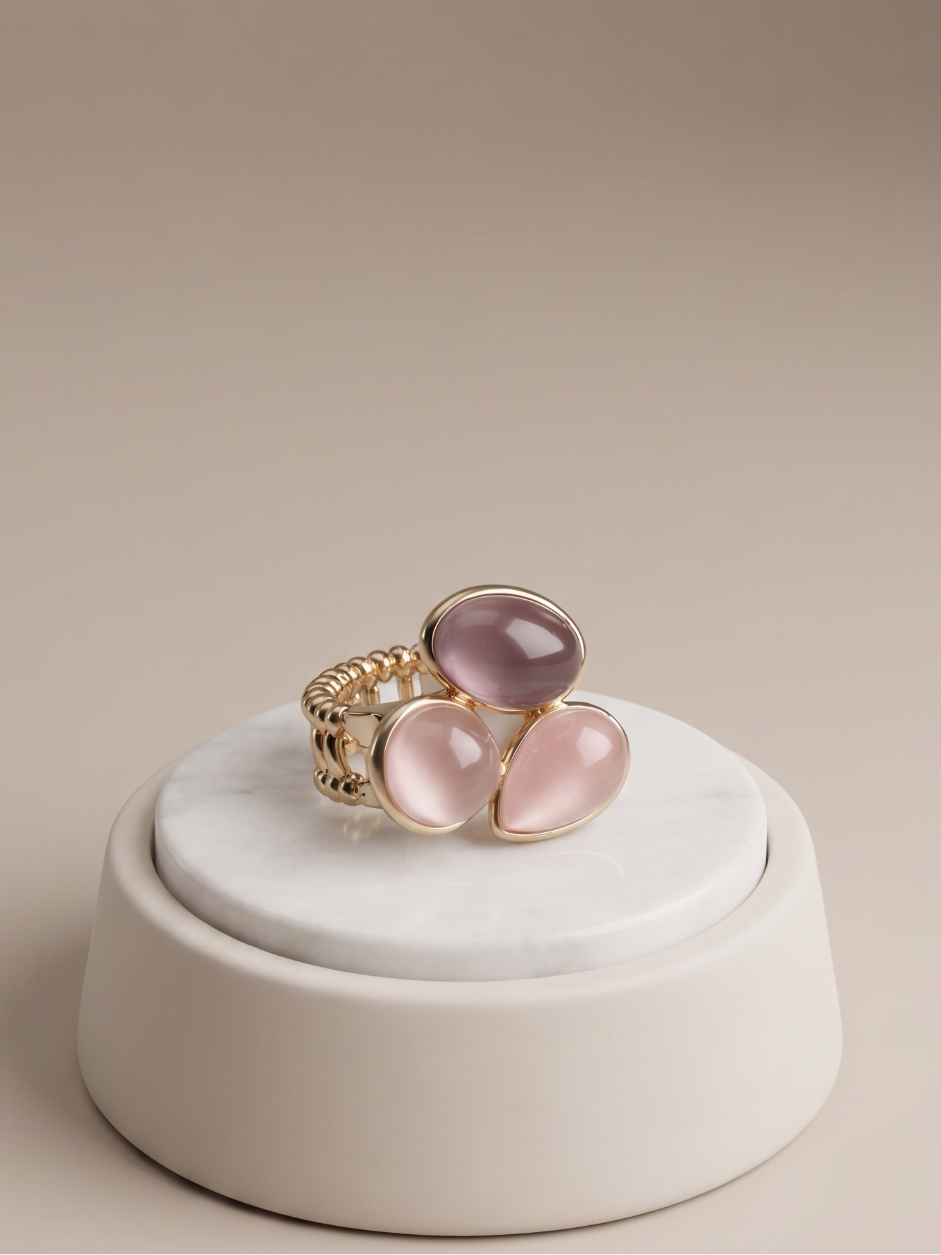 A gold ring with three oval pink cabochon gemstones, displayed on a round white marble riser against a warm beige background.
