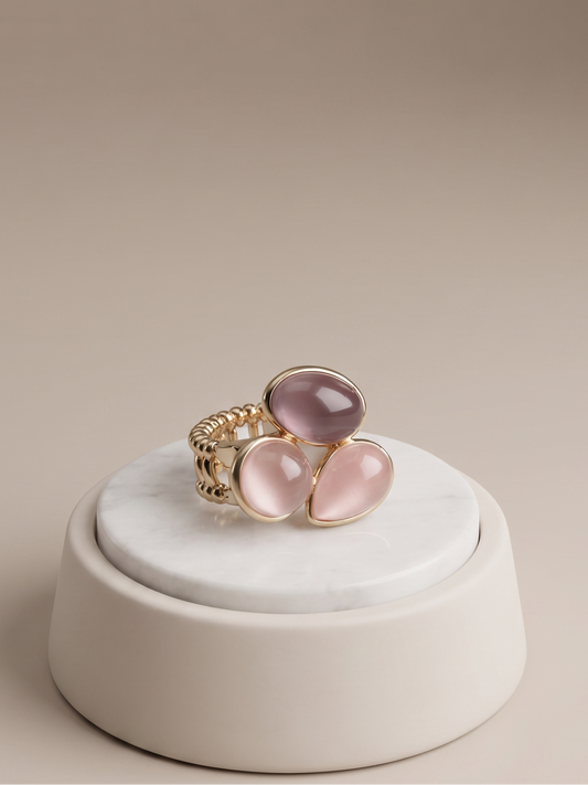 A gold ring with three oval pink cabochon gemstones, displayed on a round white marble riser against a warm beige background.