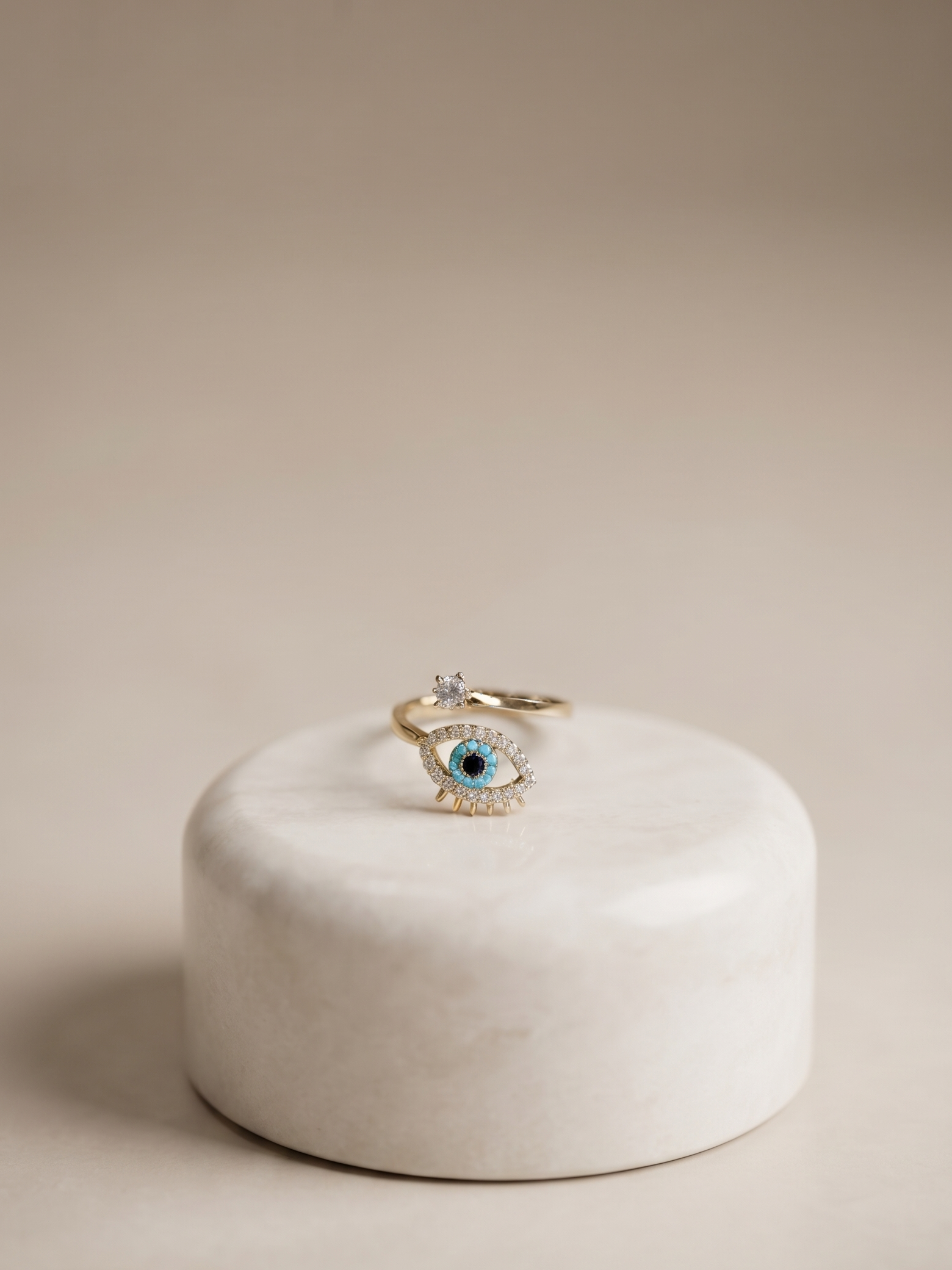 A gold-toned eye-shaped ring with a blue center and small clear stones, displayed on a round white stone pedestal against a warm beige background.