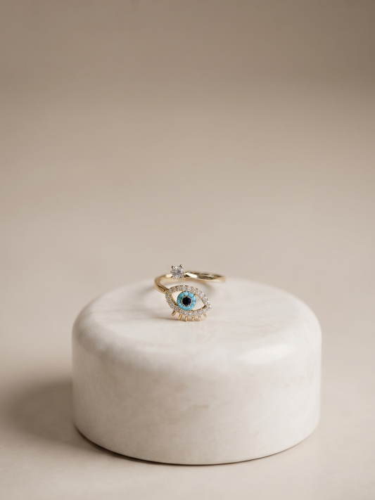 A gold-toned eye-shaped ring with a blue center and small clear stones, displayed on a round white stone pedestal against a warm beige background.