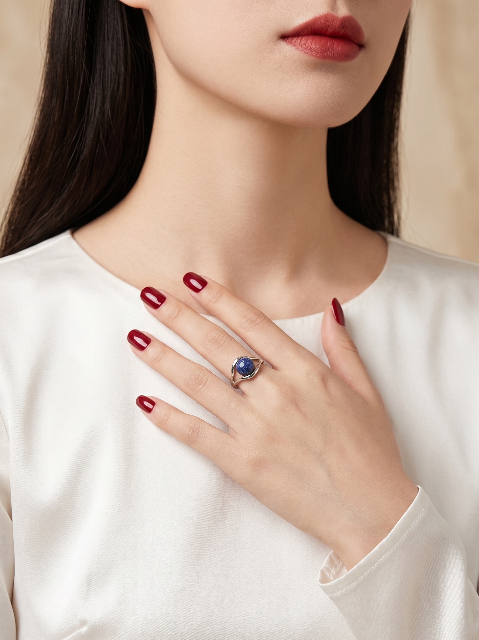 A close-up of a woman wearing a silver ring with a round blue gemstone, styled with a white satin top and dark red manicured nails.
