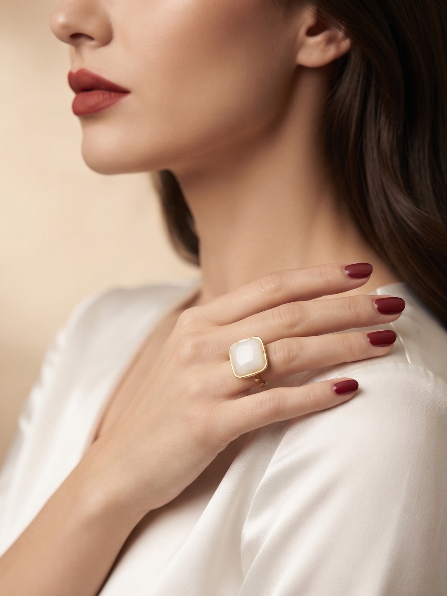 A close-up lifestyle shot of a woman wearing a gold ring with a large, rounded, milky white stone. She is wearing a white satin top, and her hand with dark red manicured nails rests on her shoulder against a warm beige background.