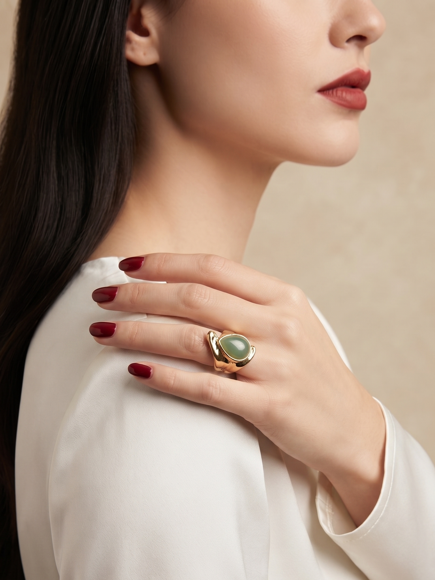 A close-up lifestyle shot of a model wearing a gold ring with a green teardrop-shaped stone, set against a soft beige background.