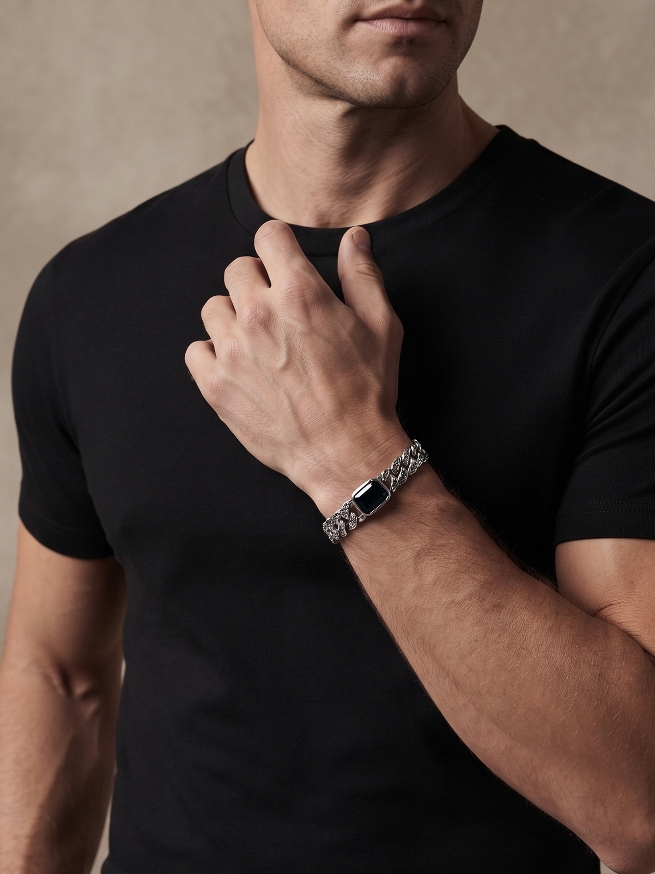 A man wearing a black short-sleeve crewneck t-shirt and black trousers, wearing a silver-toned metal link watch with a dark rectangular face on his left wrist, posed against a textured beige studio background.