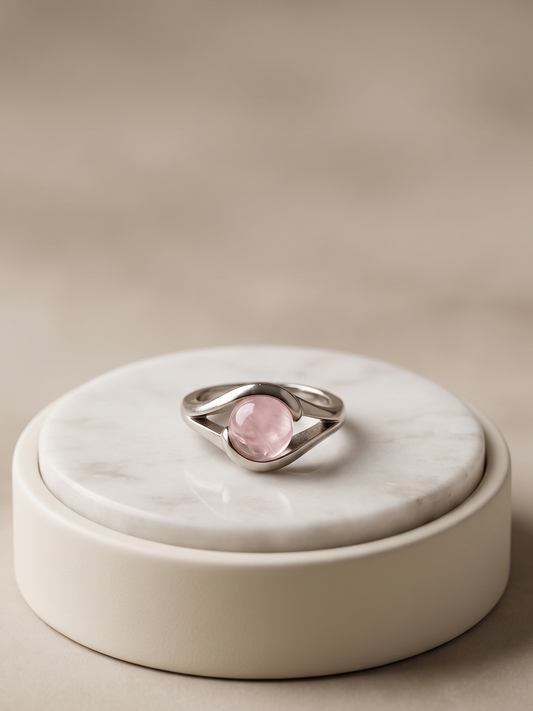 A silver ring with an oval pink gemstone, displayed on a round white marble tray set against a soft beige background.