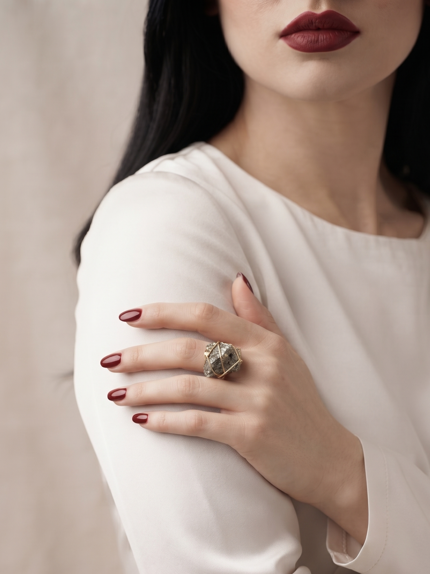 A close-up of a woman wearing a large, textured silver ring on her finger, paired with a white long-sleeve top and dark red lipstick.