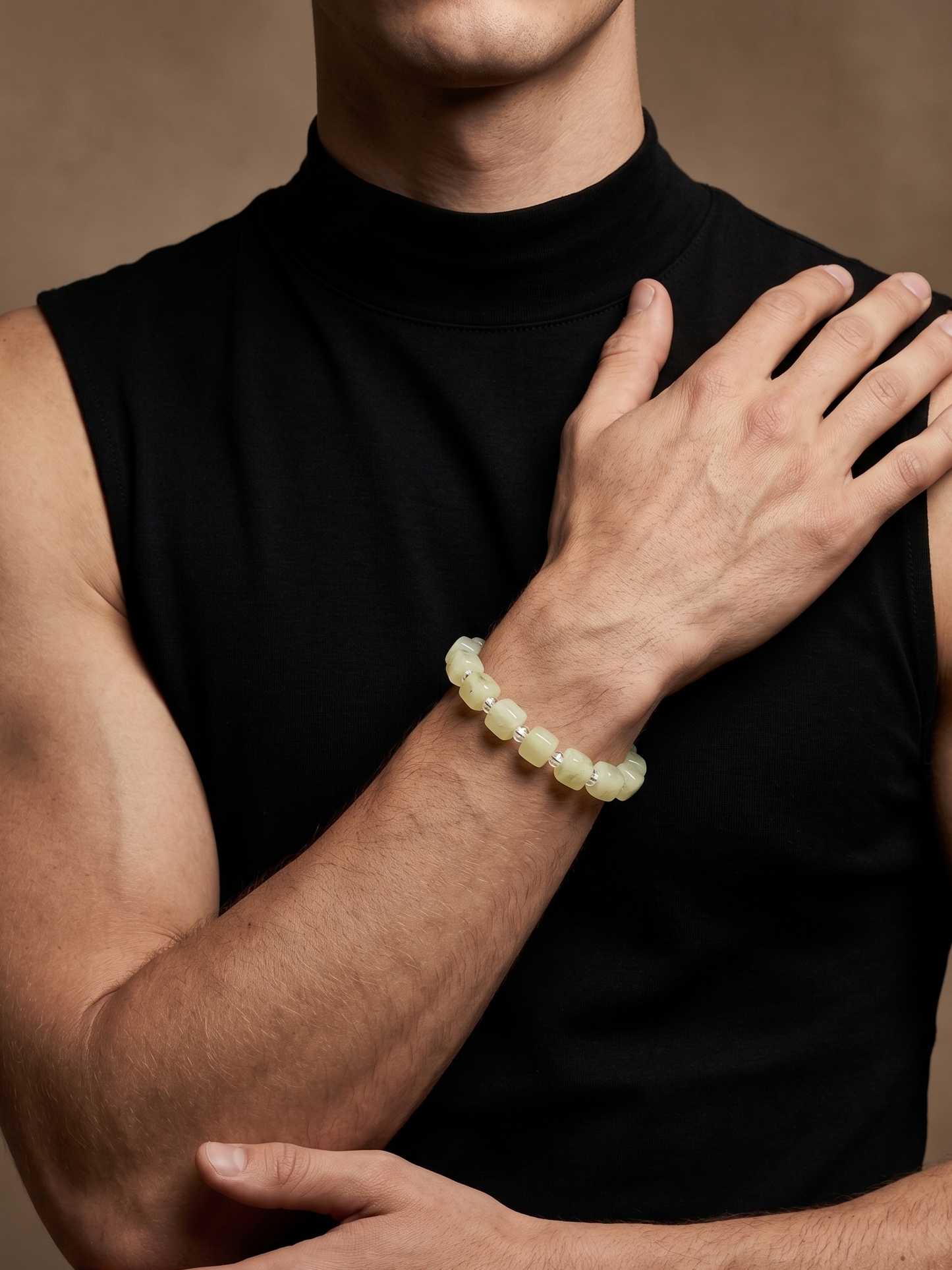 A close-up of a man wearing a pale yellow-green beaded bracelet on his wrist, wearing a black sleeveless mock-neck top against a warm brown studio background.