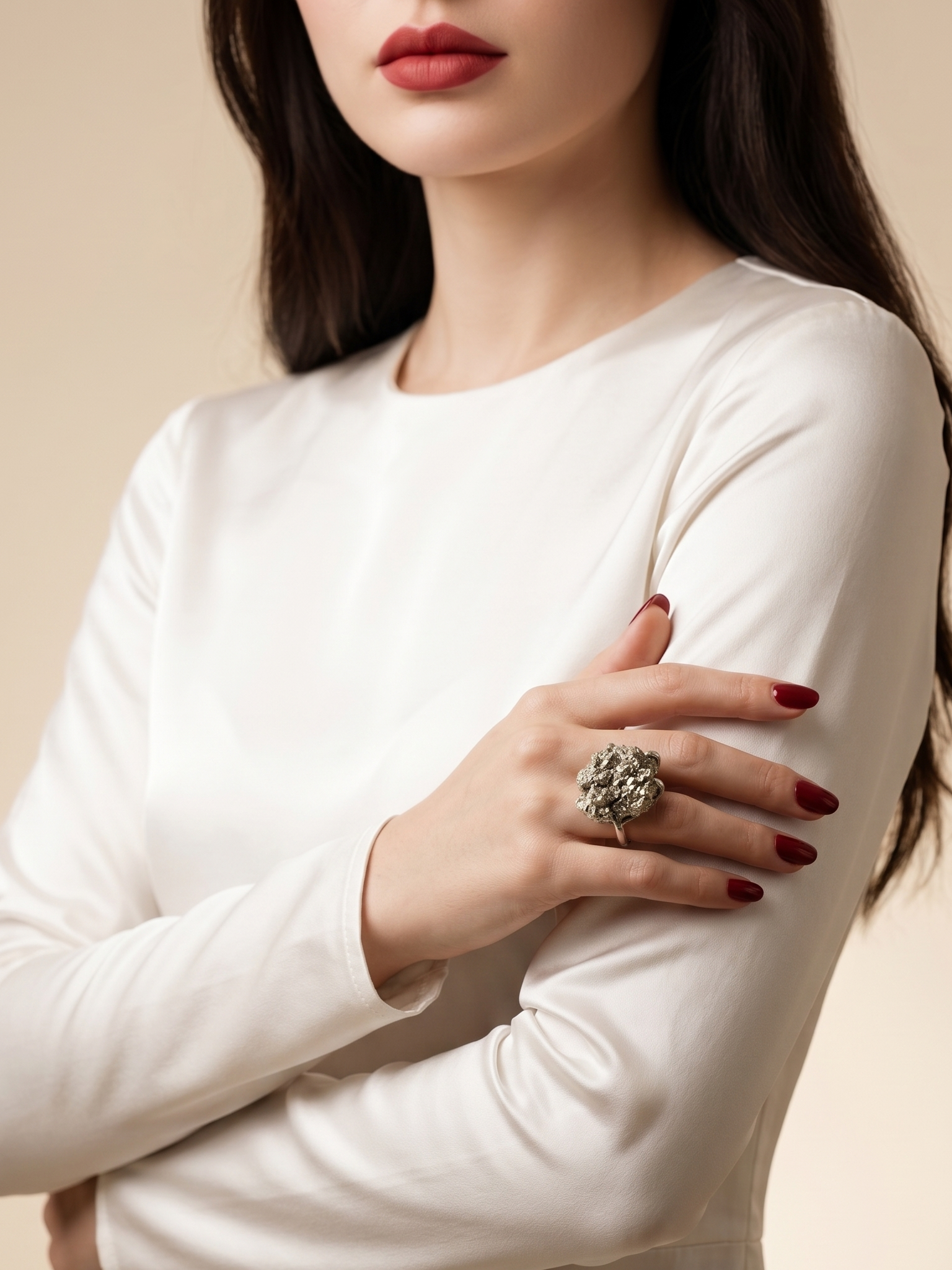A close-up of a woman wearing a cream-colored long-sleeve top and a large, textured silver ring on her hand, posed against a solid beige background.