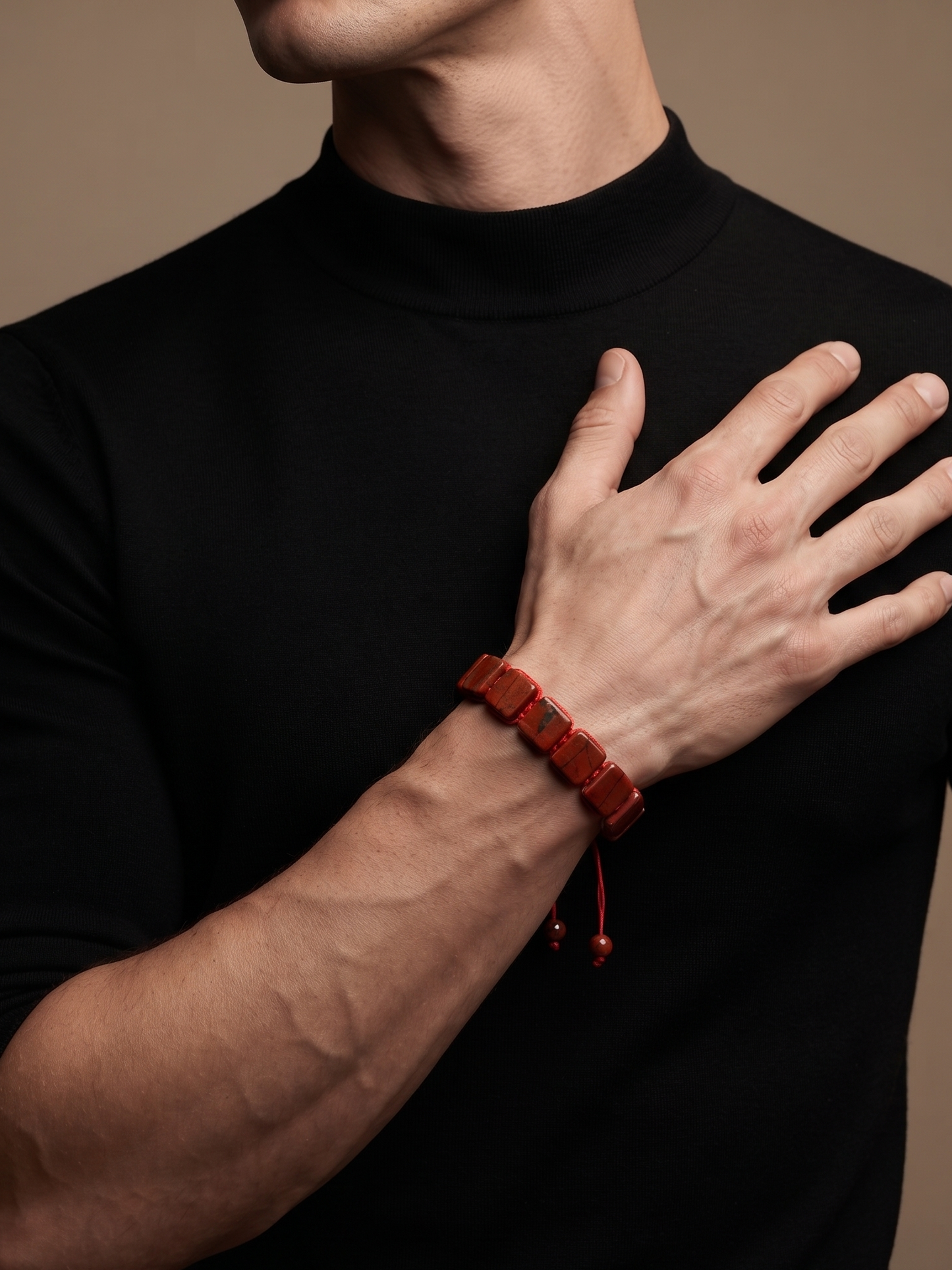 A close-up of a man wearing a black mock-neck t-shirt and a dark red beaded bracelet on his wrist, set against a solid warm brown background.
