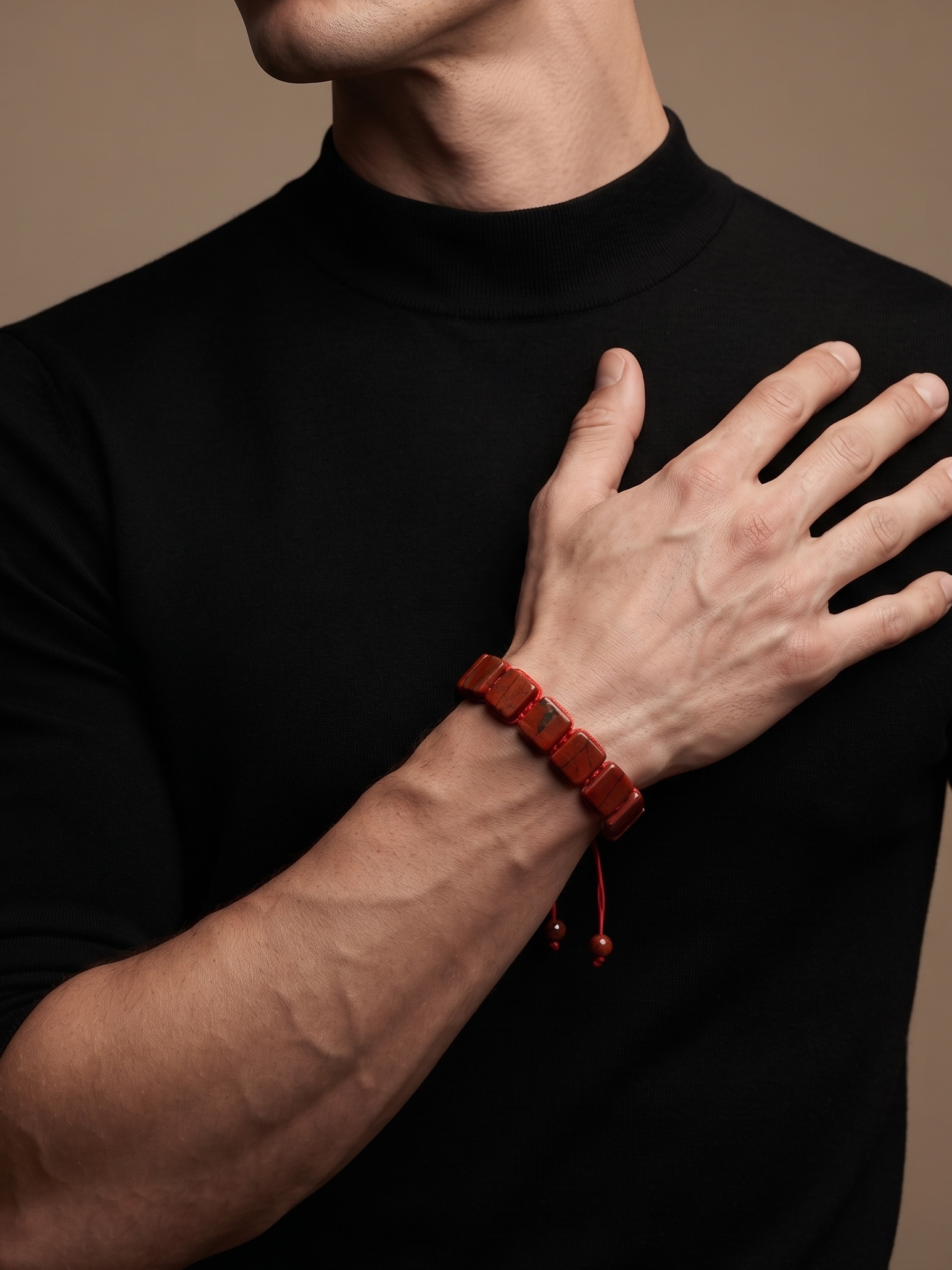 A close-up of a man wearing a black mock-neck t-shirt and a dark red beaded bracelet on his wrist, set against a solid warm brown background.