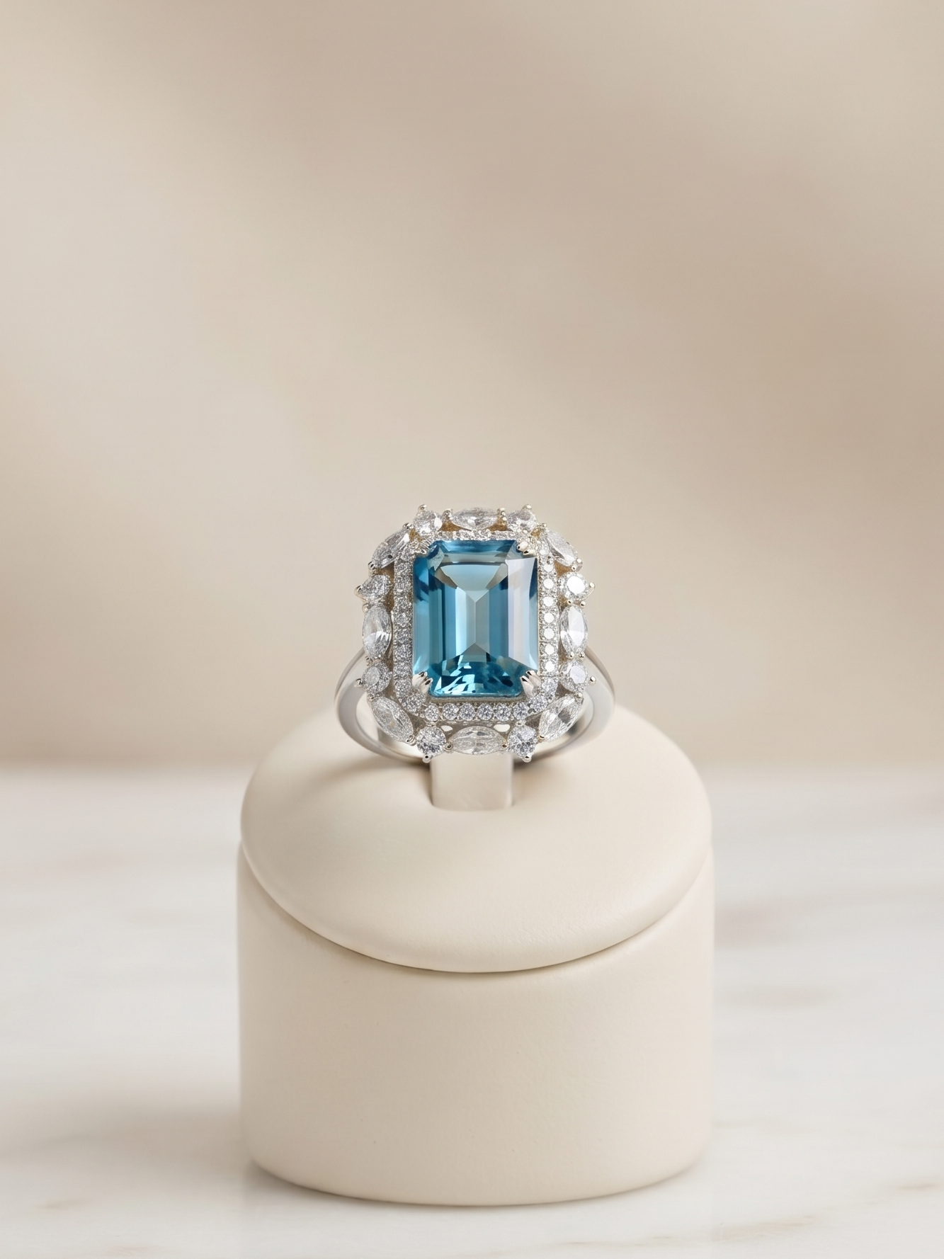 A silver ring featuring a large emerald-cut blue gemstone surrounded by smaller clear stones, displayed on a cream-colored ring stand over a light marble surface.