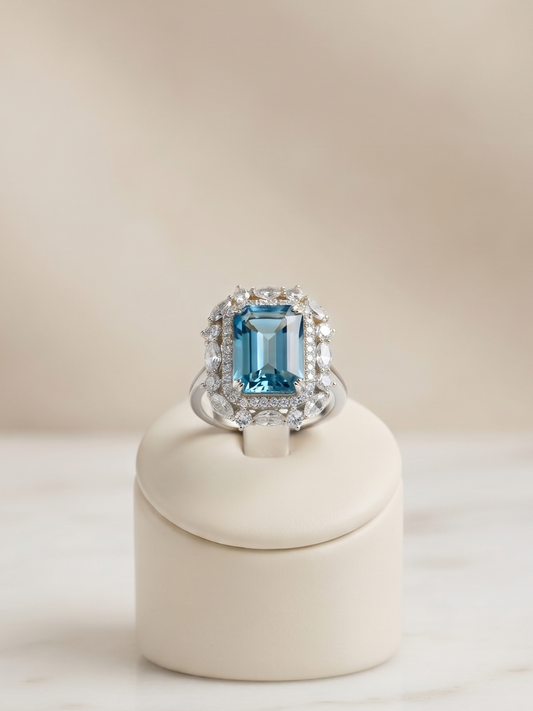 A silver ring featuring a large emerald-cut blue gemstone surrounded by smaller clear stones, displayed on a cream-colored ring stand over a light marble surface.