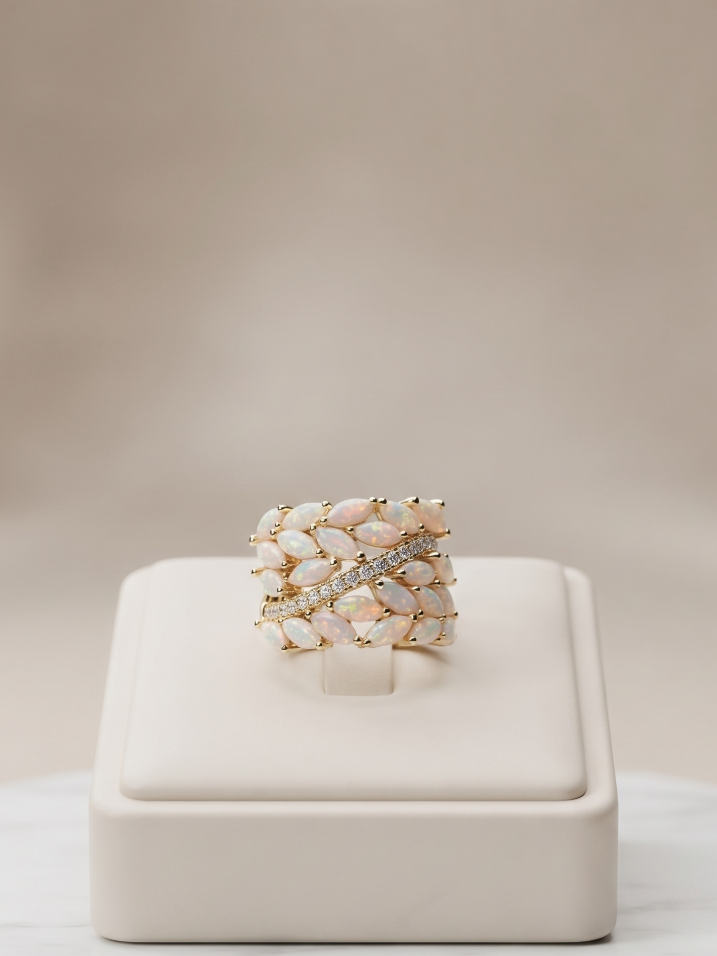 A wide band ring with a textured, woven pattern of pale pink and cream stones, accented by a central clear crystal, displayed on a cream jewelry stand over a white marble surface.