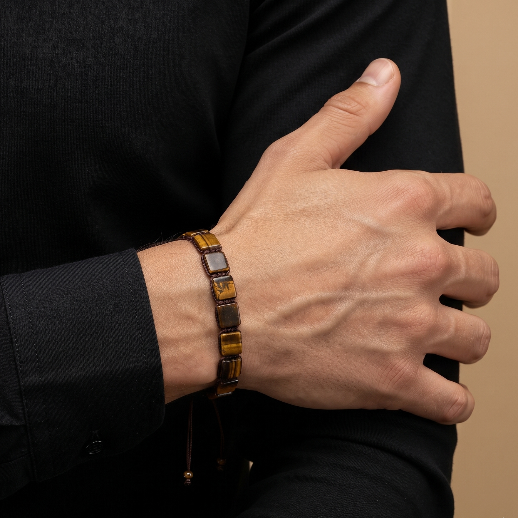 A close-up of a person's hand and forearm wearing a brown leather bracelet with polished gold-toned rectangular links and dark square accents, set against a black shirt and a warm beige background.
