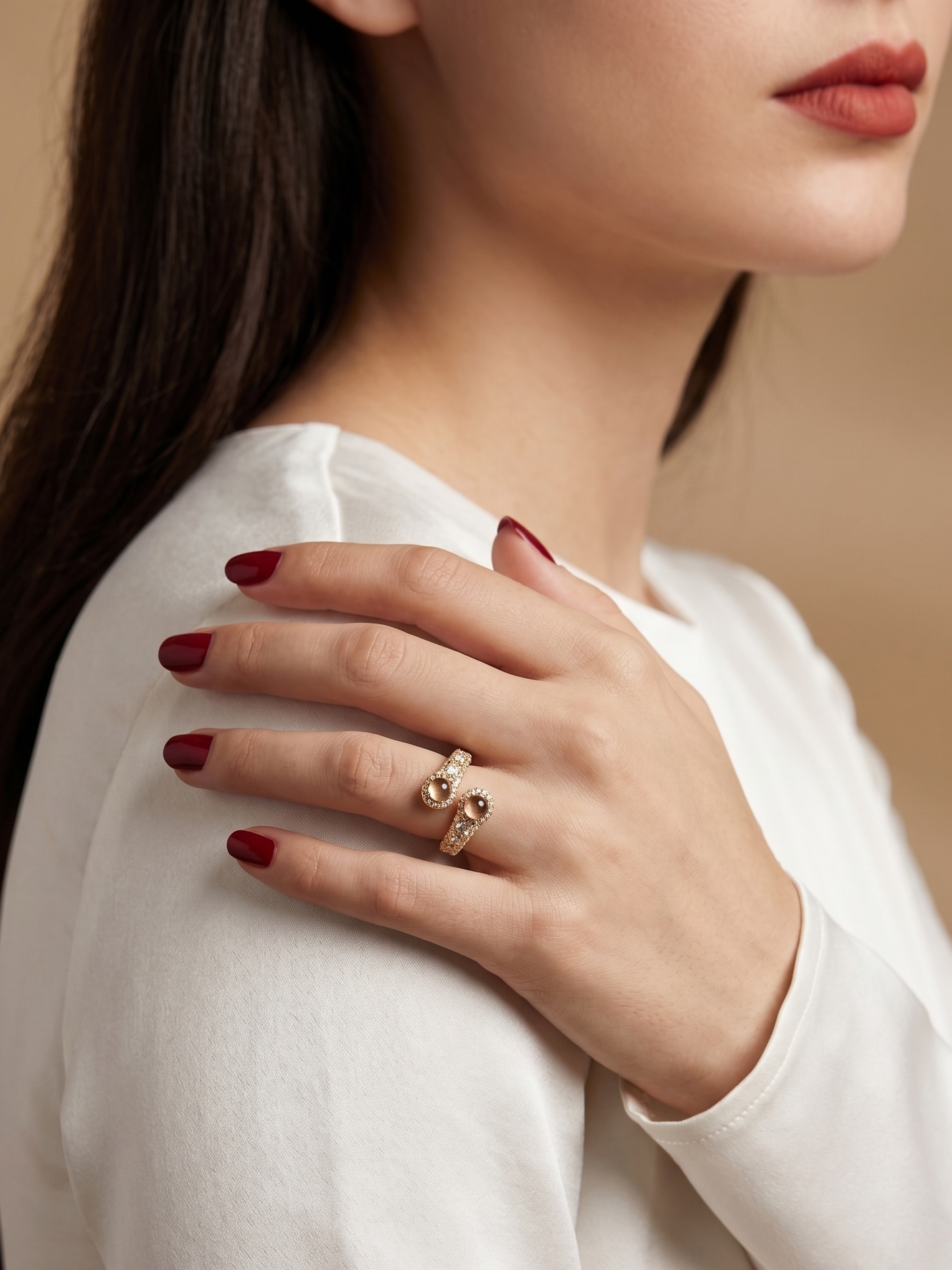 A close-up lifestyle shot of a woman wearing a gold snake ring on her finger, with her hand resting on her shoulder. She is wearing a white long-sleeve top and has dark red nail polish against a warm beige background.