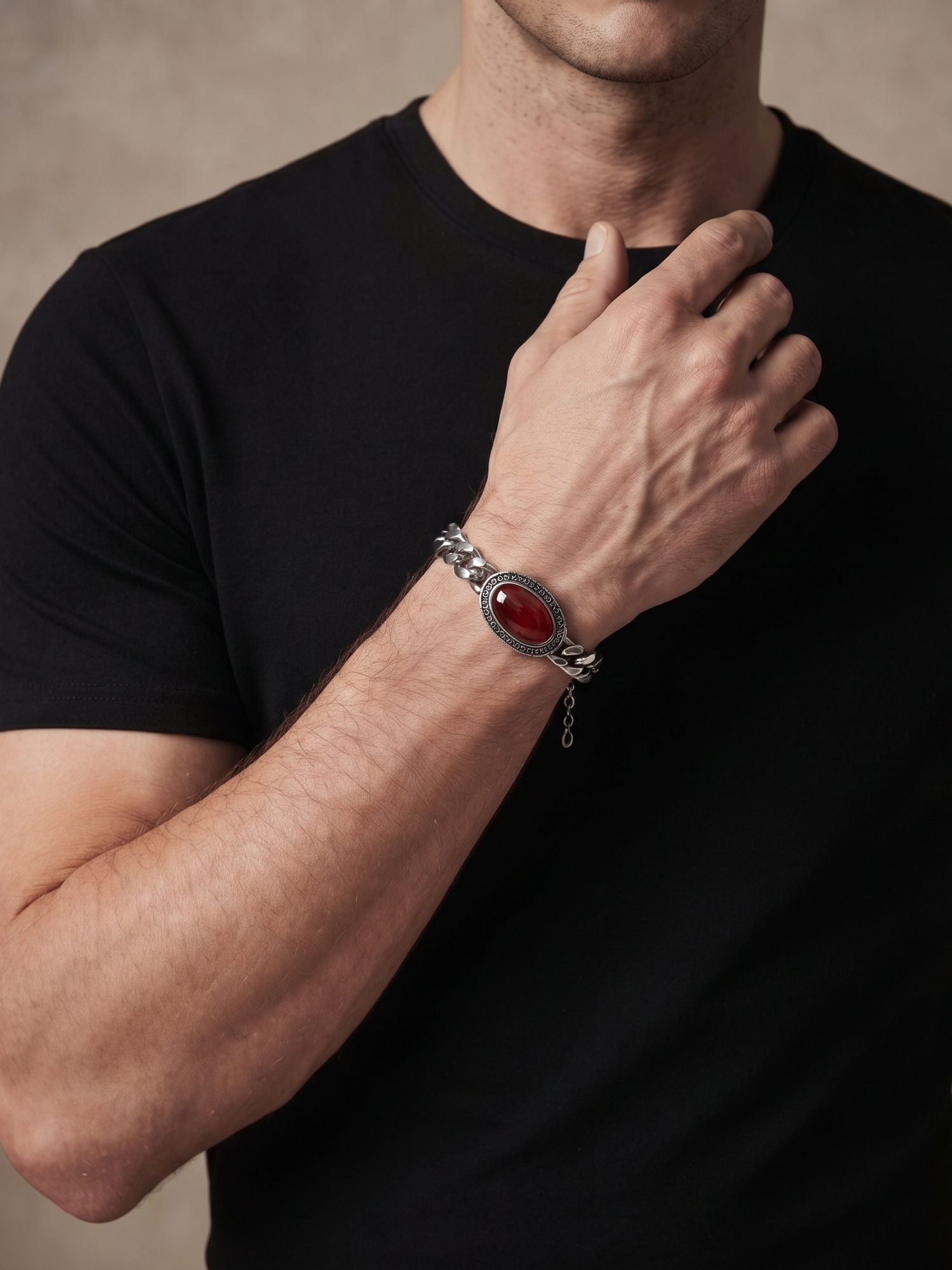 A close-up of a man wearing a black t-shirt and black trousers, showcasing a silver-toned chain bracelet with a prominent oval red cabochon stone on his wrist, set against a neutral textured beige background.
