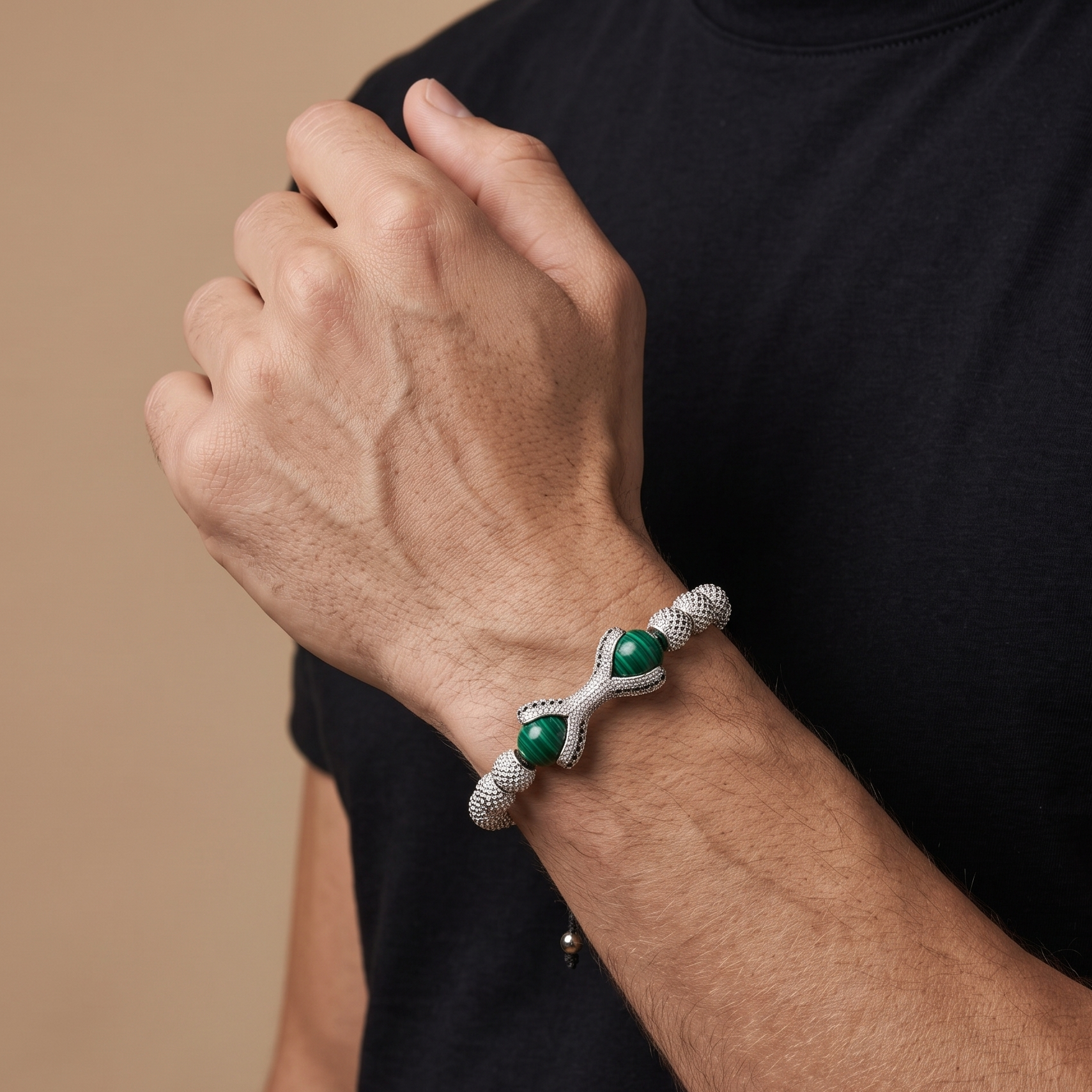 A close-up of a man's wrist wearing a silver-toned textured bracelet with two green gemstone accents, set against a black t-shirt and a warm beige background.