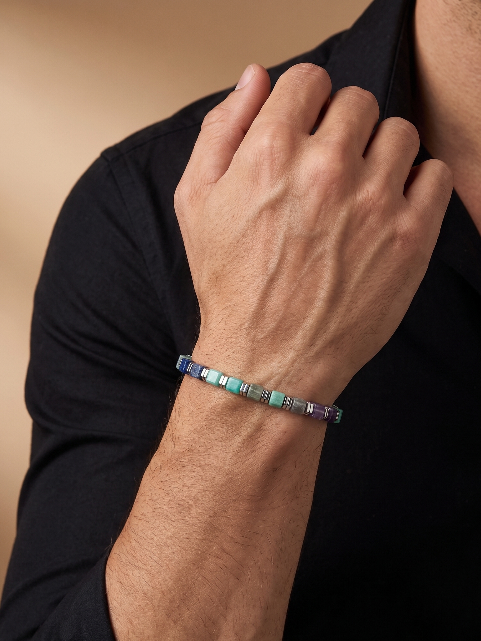 A close-up of a person's hand and wrist wearing a beaded bracelet with iridescent square beads in shades of blue, green, and purple, worn over a black shirt.