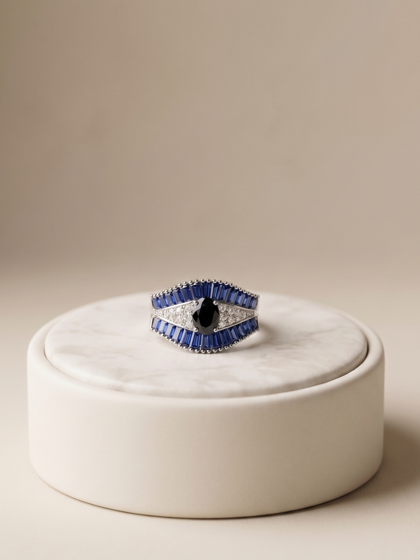 A geometric silver ring with blue and white textured inlay, featuring a central black heart-shaped stone, displayed on a round white marble pedestal against a warm beige background.