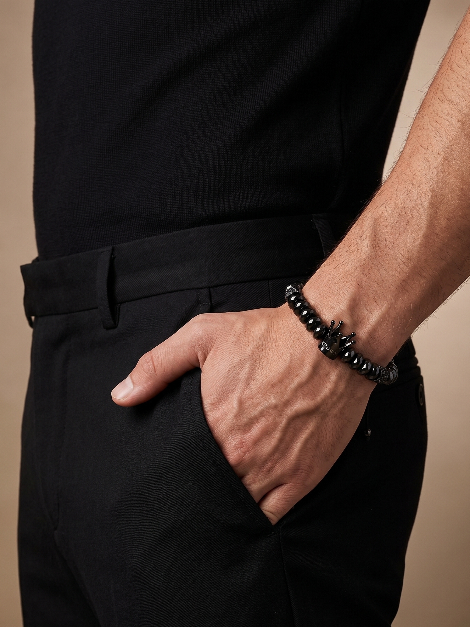 A close-up of a man's hand resting in his pocket, wearing a black beaded bracelet with a silver-toned anchor charm, paired with black trousers and a black shirt against a warm beige background.