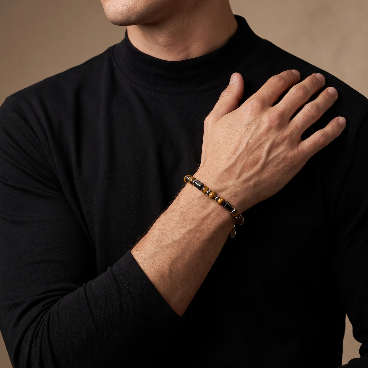 A close-up of a man wearing a black long-sleeve mock neck shirt and a beaded bracelet on his wrist, with his hand resting on his shoulder against a warm beige background.