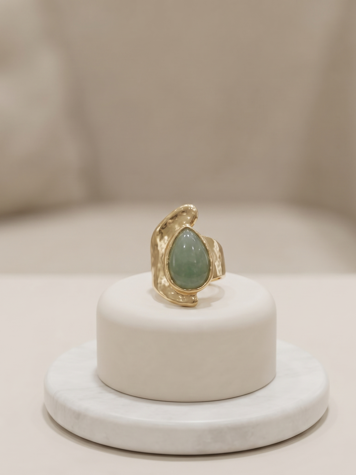 A gold ring with a teardrop-shaped light green stone, displayed on a small white cylindrical pedestal.