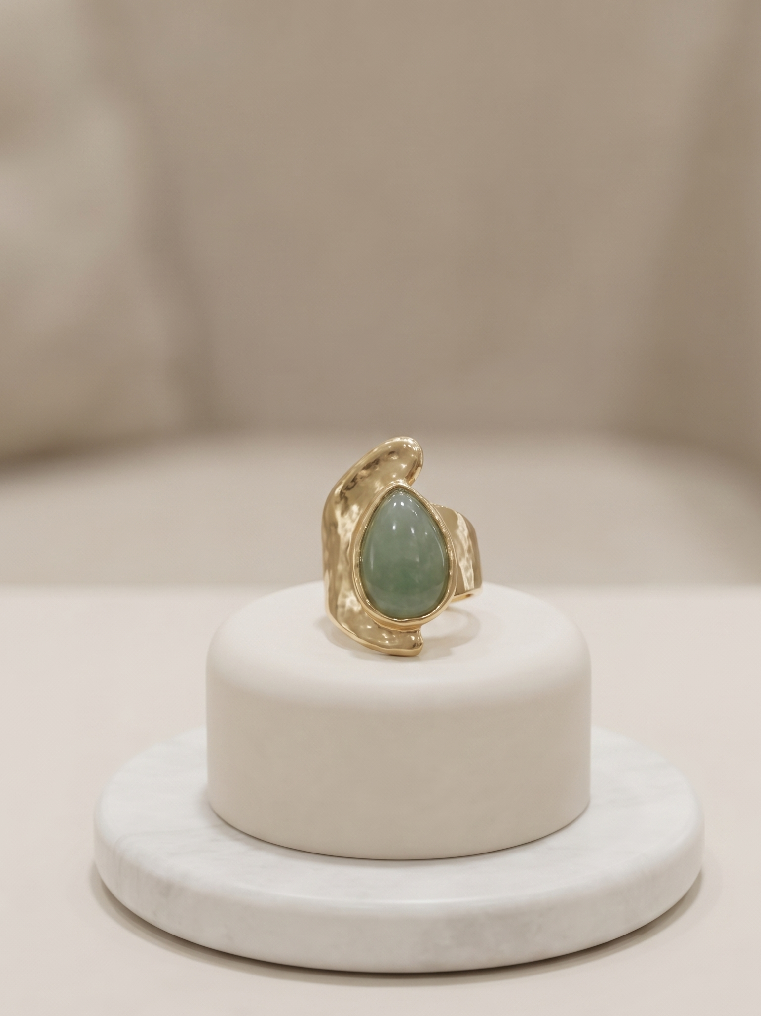 A gold ring with a teardrop-shaped light green stone, displayed on a small white cylindrical pedestal.