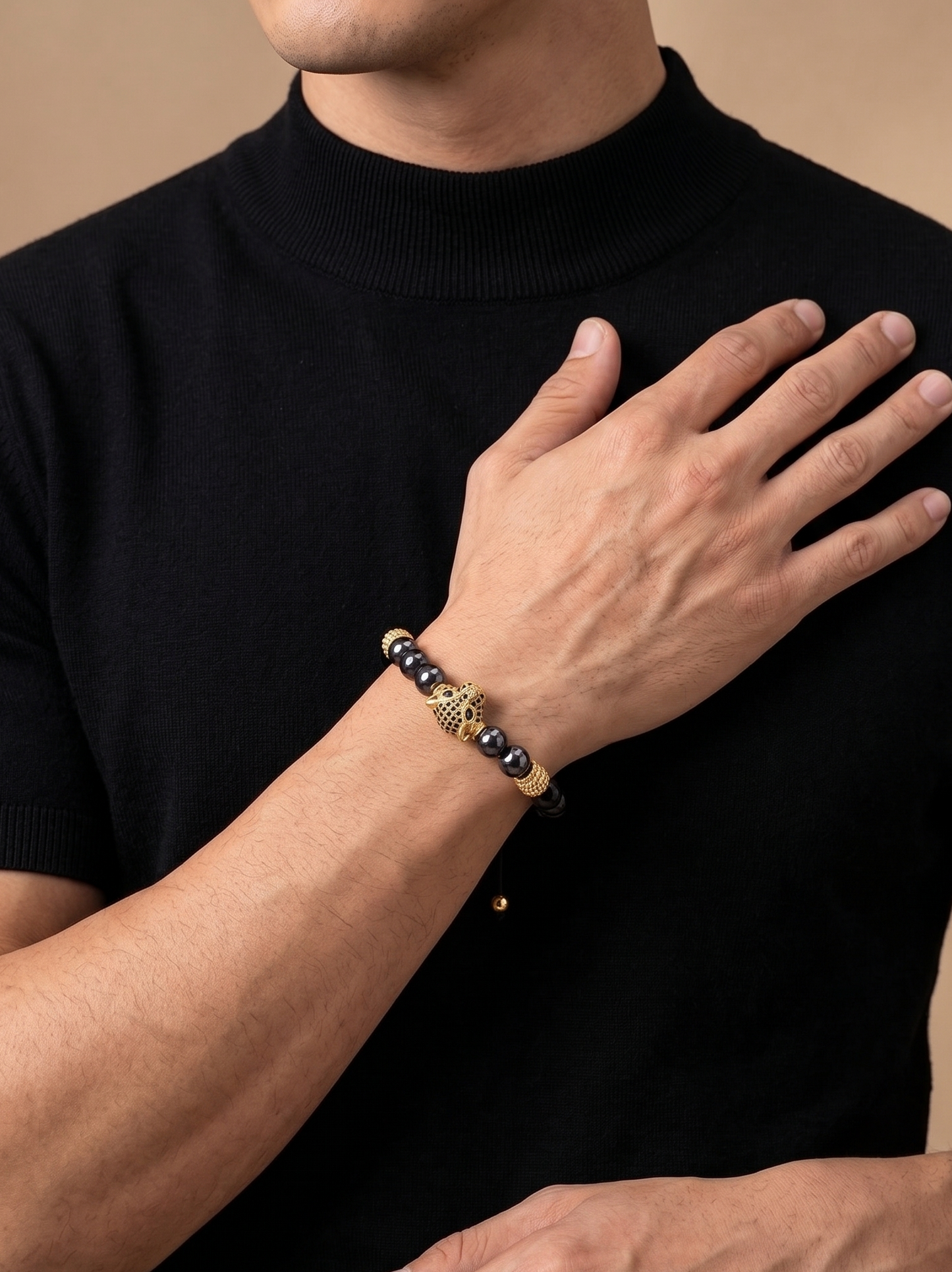 A close-up of a man wearing a black turtleneck sweater, showcasing a beaded bracelet on his wrist. The bracelet features dark gray beads, a central gold-toned elephant charm, and a thin black cord.