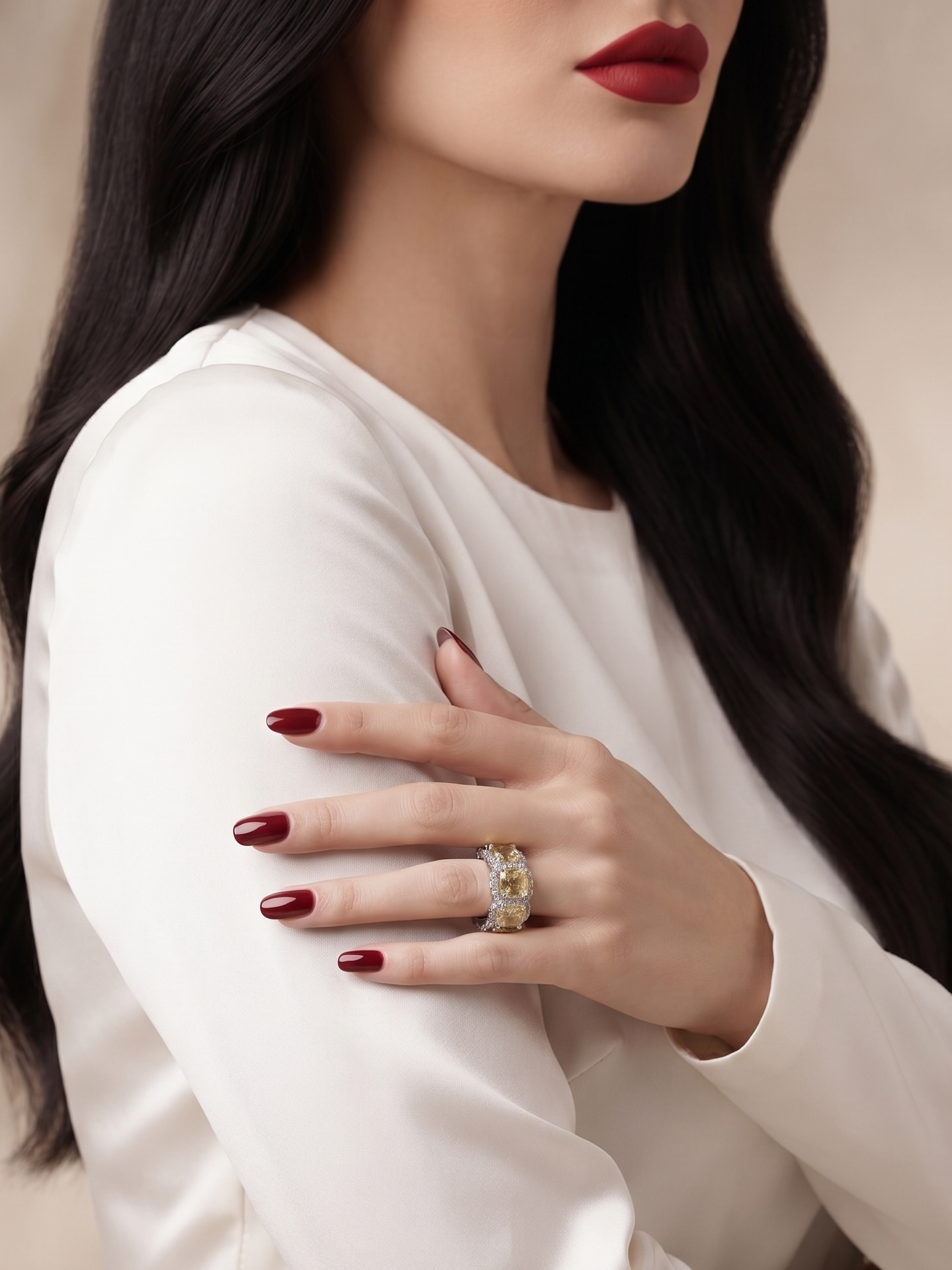 A close-up of a woman wearing a white satin long-sleeve top and a statement ring on her finger, with her hand resting on her arm against a soft beige background.