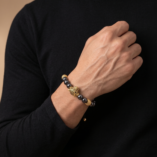 A close-up of a man's wrist wearing a beaded bracelet with dark gray beads, a gold-toned lion head charm, and a small gold accent bead, styled with a black long-sleeve shirt against a warm beige background.