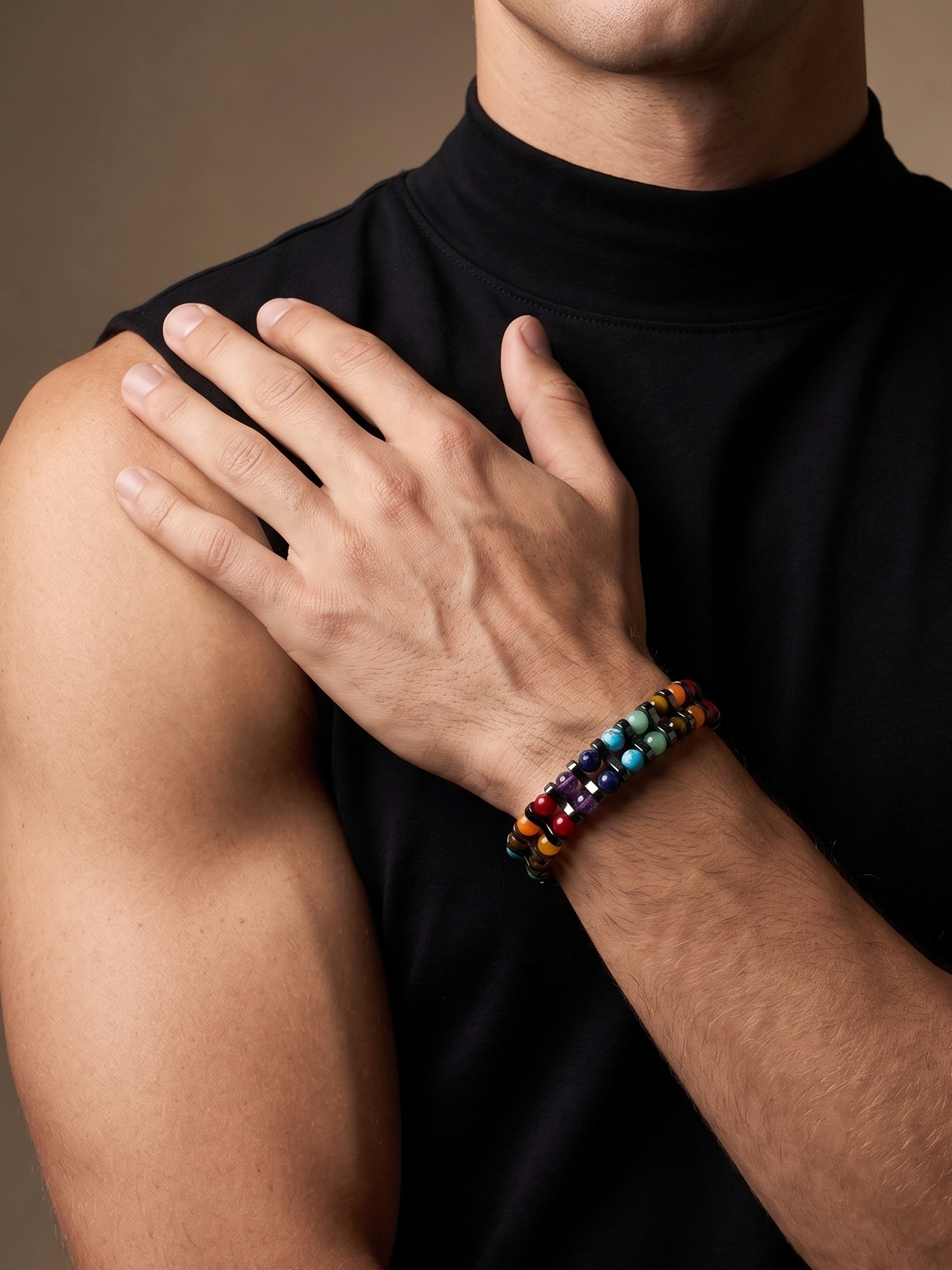 A close-up of a man wearing a beaded stretch bracelet on his wrist, showing the colorful beads. He is wearing a black sleeveless t-shirt against a warm brown studio background.