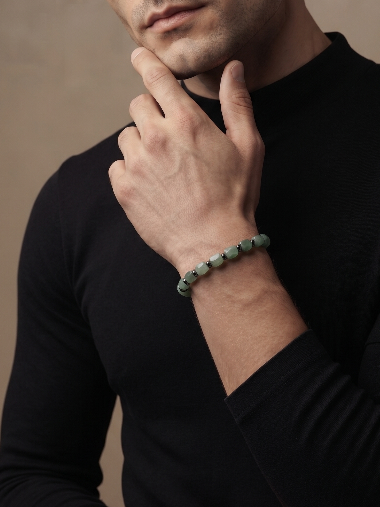 A man wearing a black long-sleeve mock neck shirt and a green beaded bracelet, posing with his hand on his chin against a textured beige background.