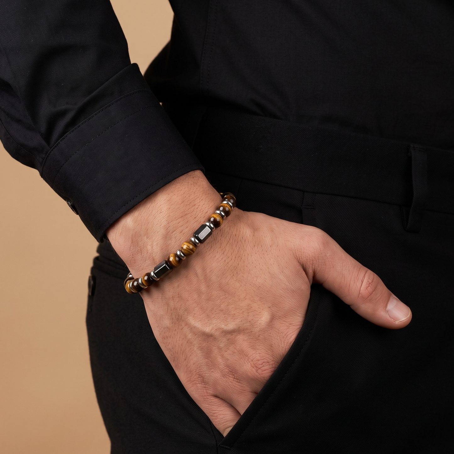 A close-up of a man's hand in his pocket wearing a beaded bracelet with brown, black, and metallic silver beads, dressed in a black shirt and trousers against a warm beige background.