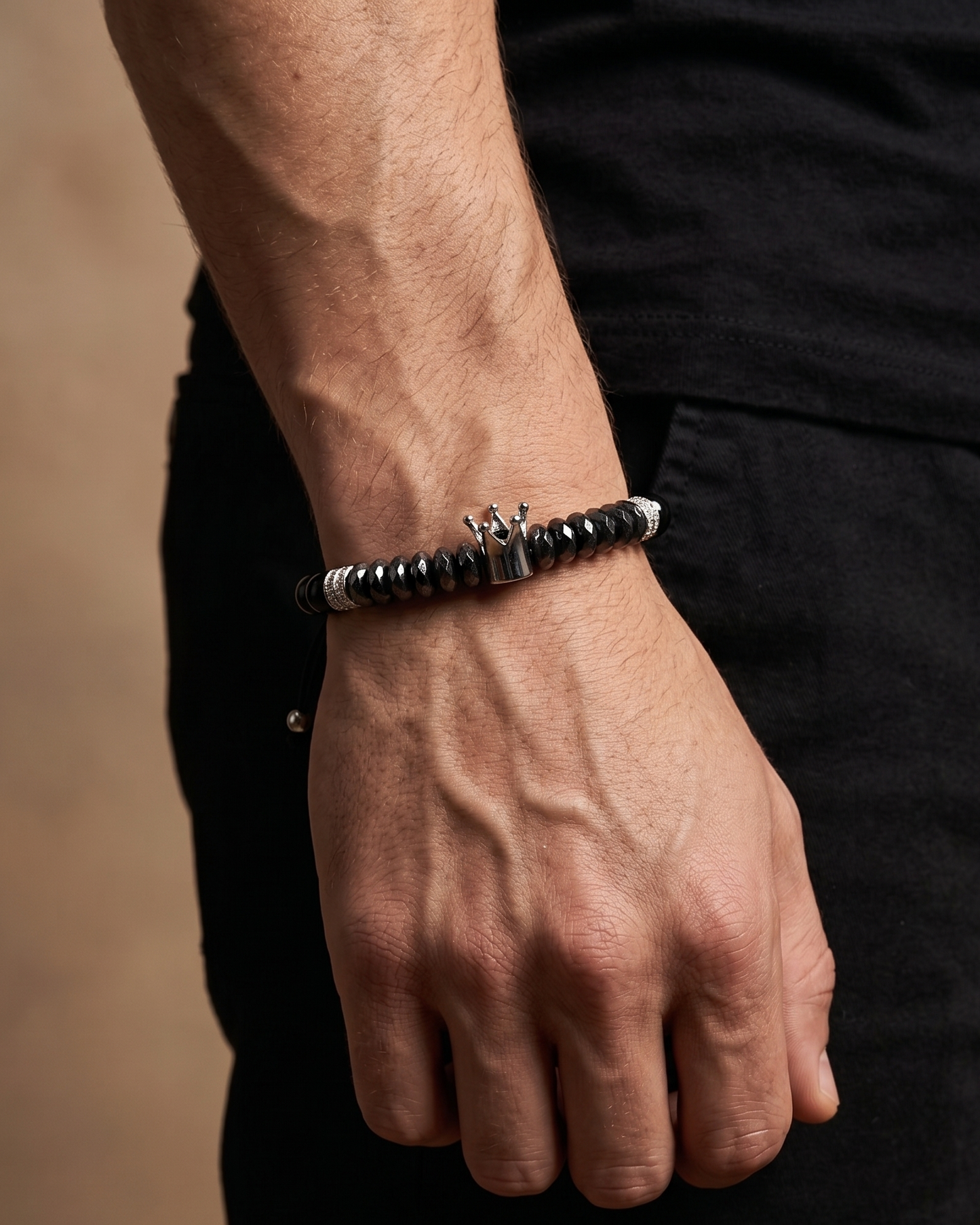 A close-up of a man's hand and wrist wearing a black beaded bracelet with a silver-toned metal centerpiece, paired with a black shirt against a warm beige background.