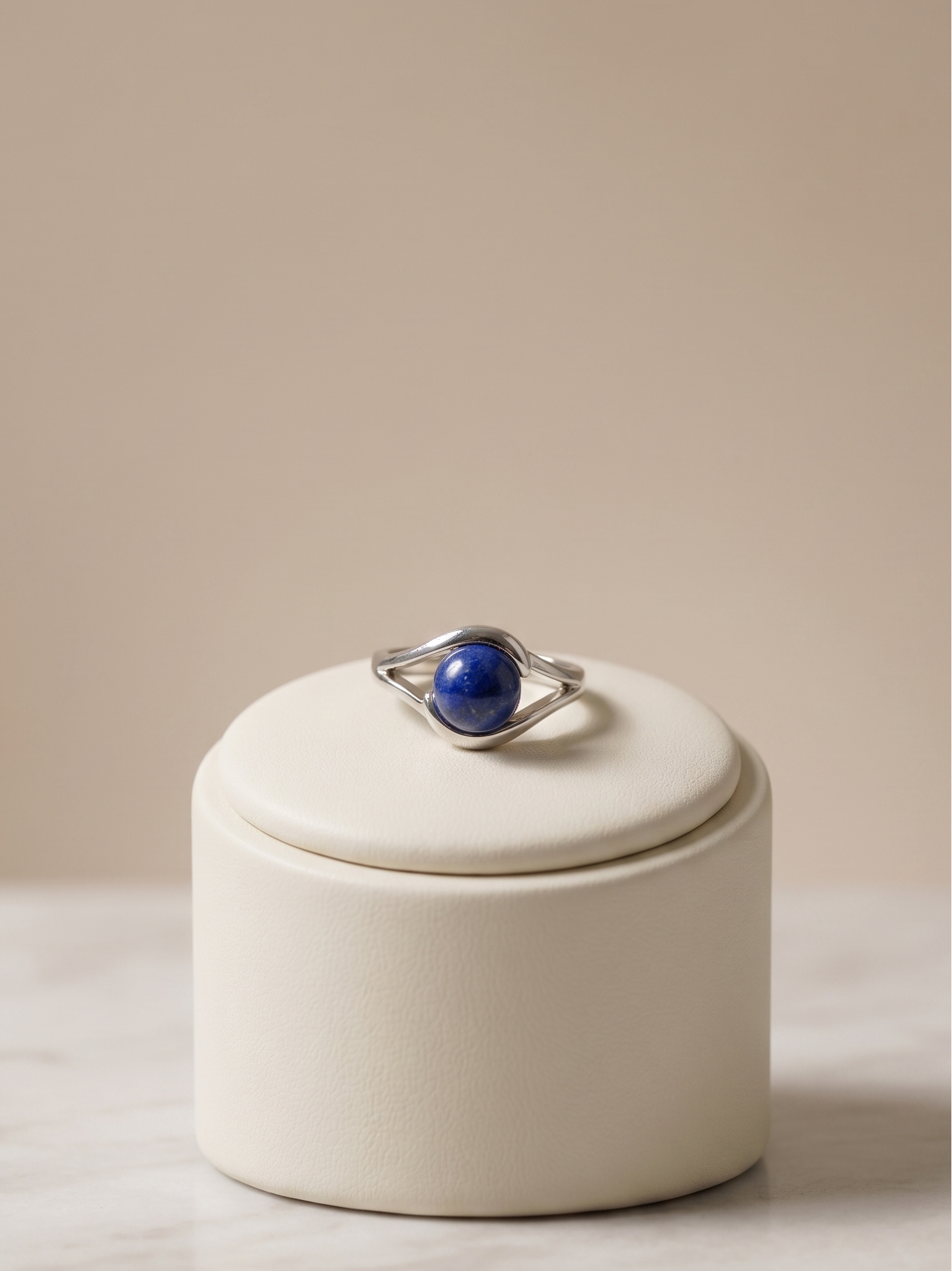 A silver ring with a polished blue oval stone, displayed on the lid of a cream-colored cylindrical jewelry box on a light marble surface against a warm beige background.