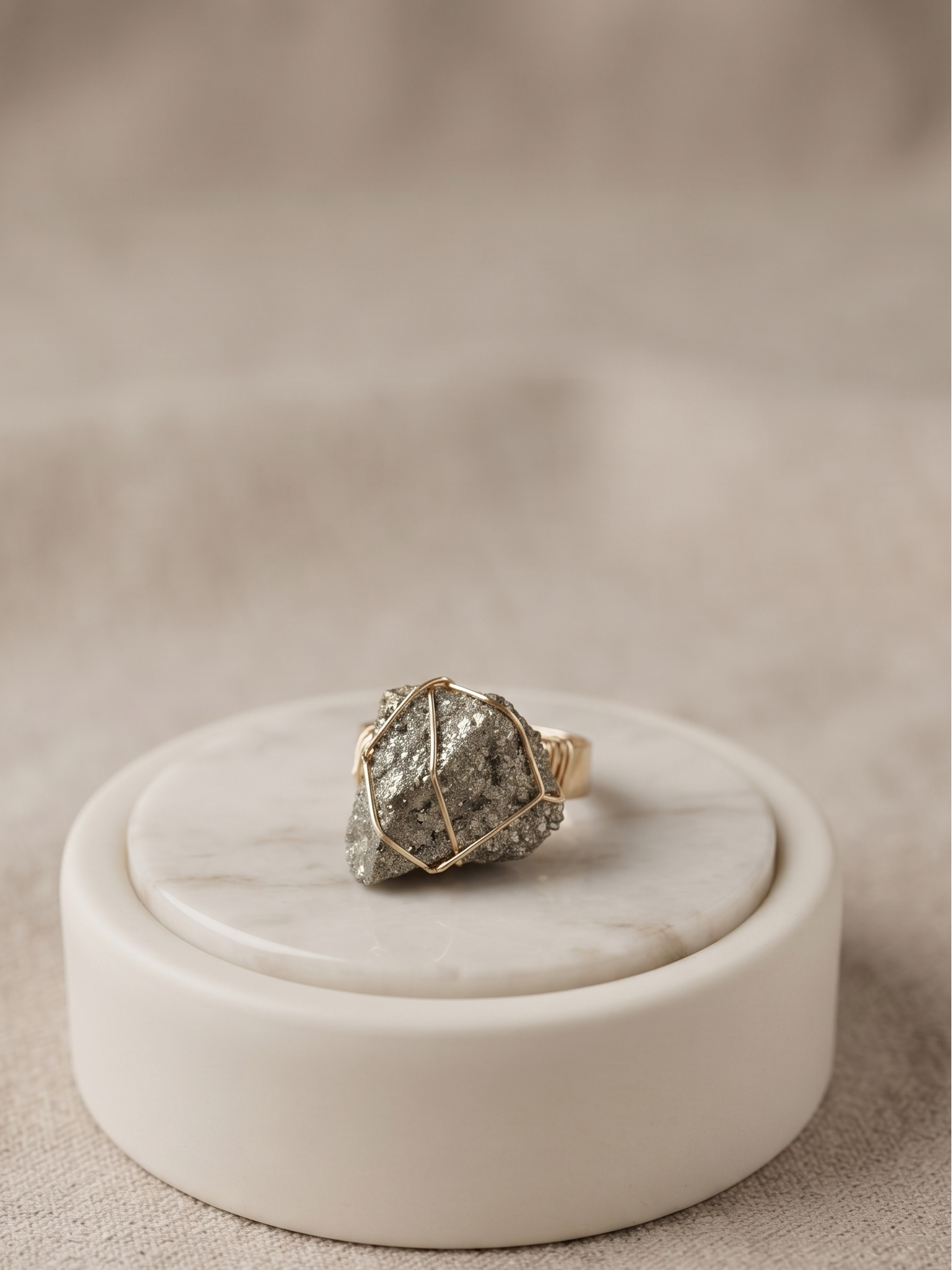 A gold ring with a textured, rough stone featuring gray and white marbling, displayed on a round white marble stand against a soft beige fabric background.