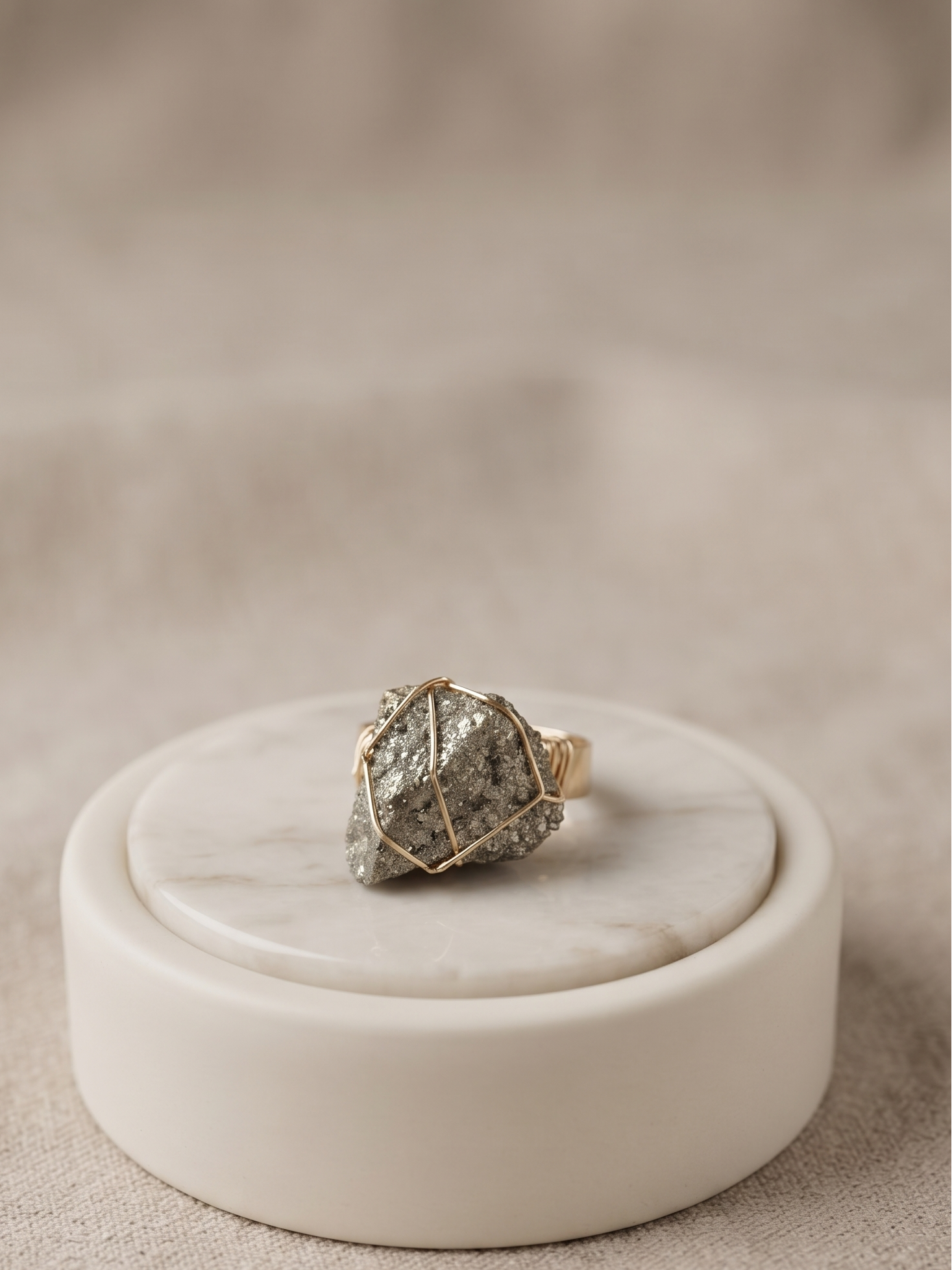 A gold ring with a textured, rough stone featuring gray and white marbling, displayed on a round white marble stand against a soft beige fabric background.