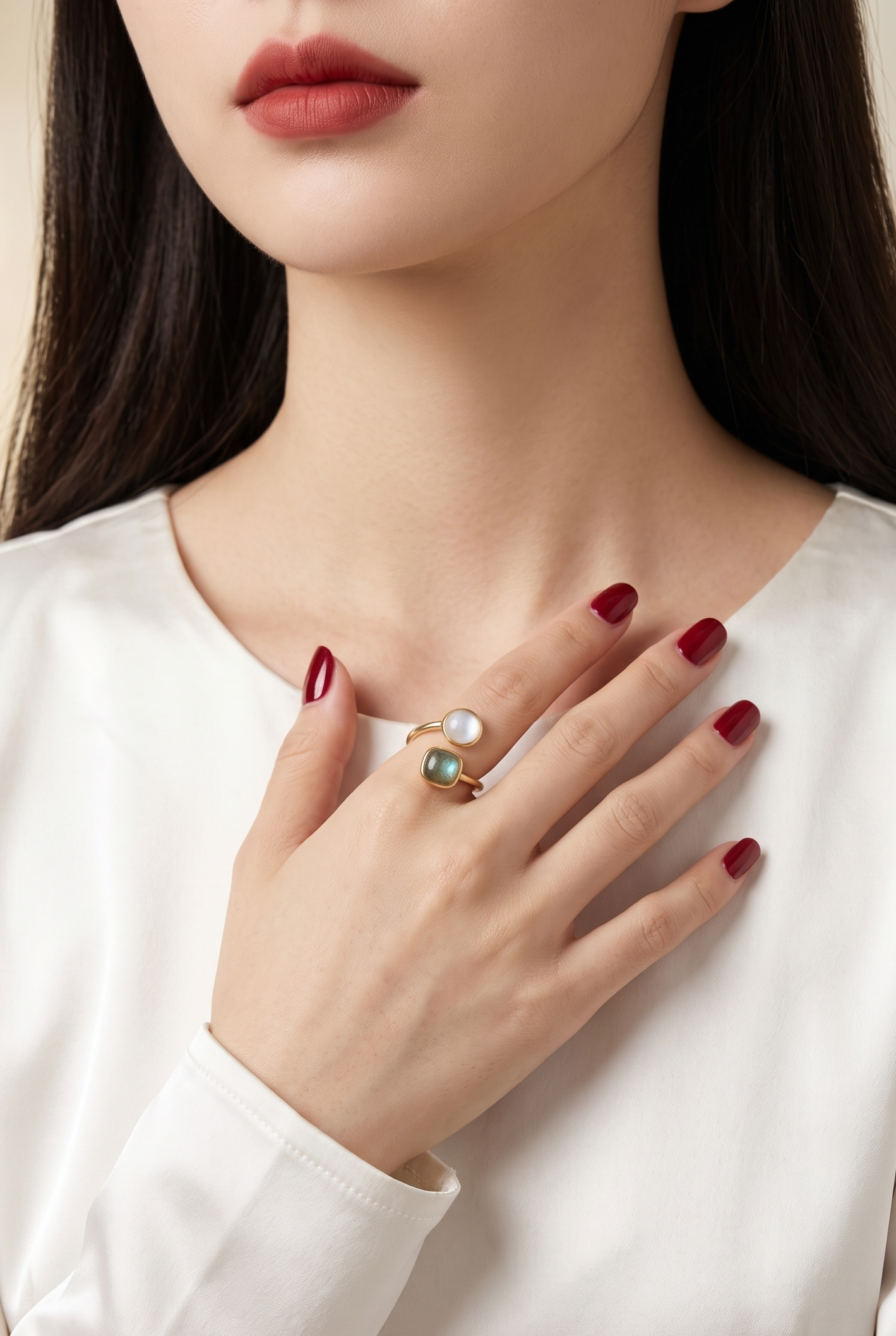 A close-up of a woman wearing a gold ring with a white pearl and a green stone, styled on her hand as she wears a white satin top.