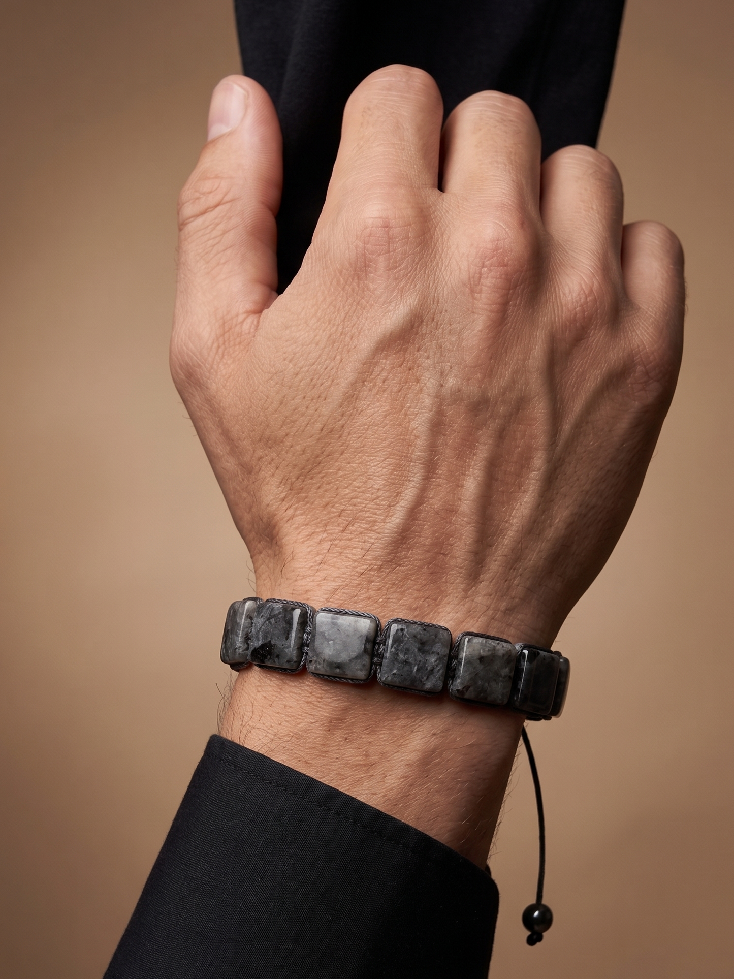 A close-up of a person's hand wearing a dark gray stone bracelet with a black cord, shown against a warm brown background.