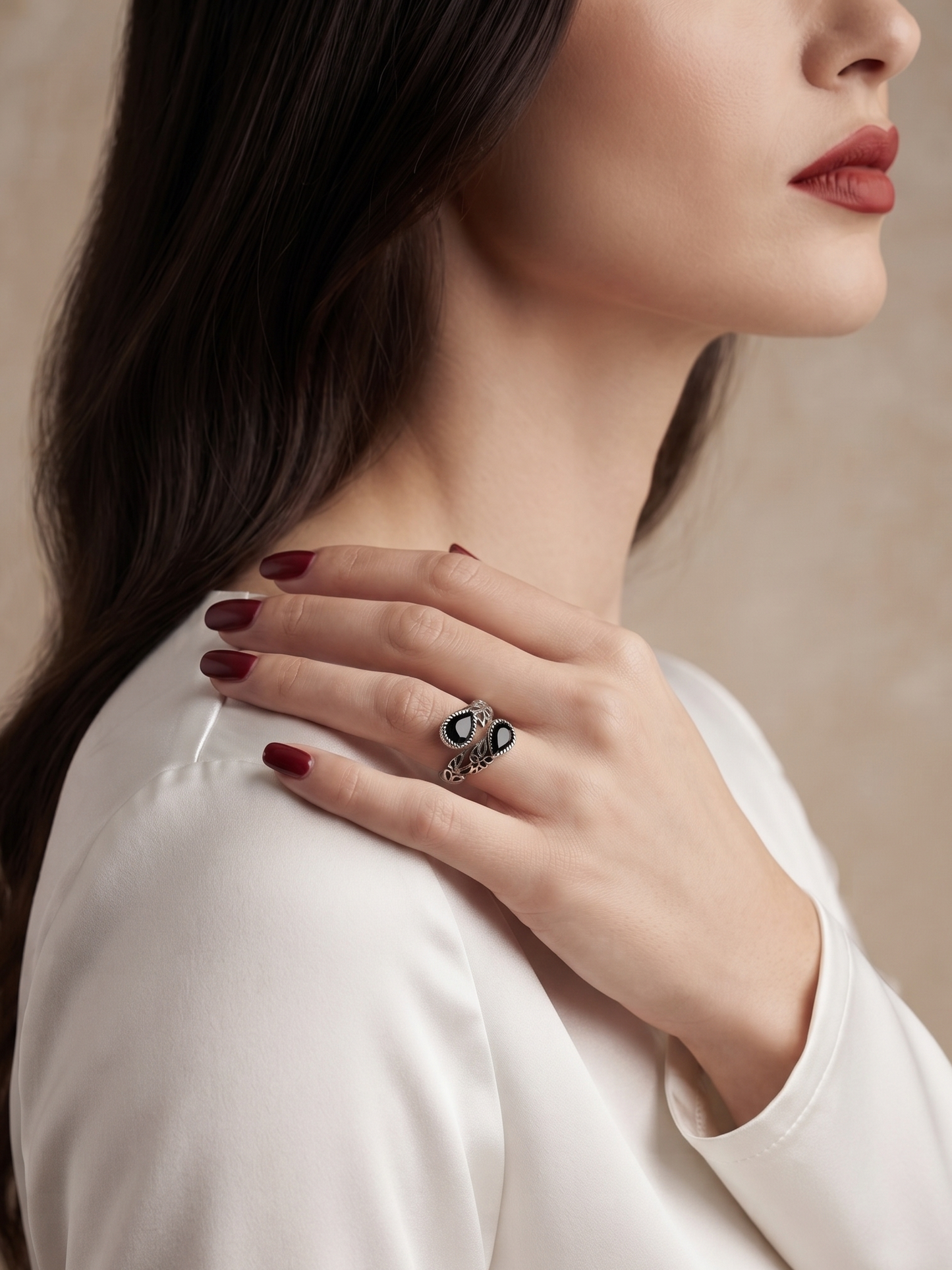 A close-up lifestyle shot of a woman wearing a silver ring with black and white enamel details on her finger, with her hand resting on her shoulder. She is wearing a white satin blouse against a neutral beige background.