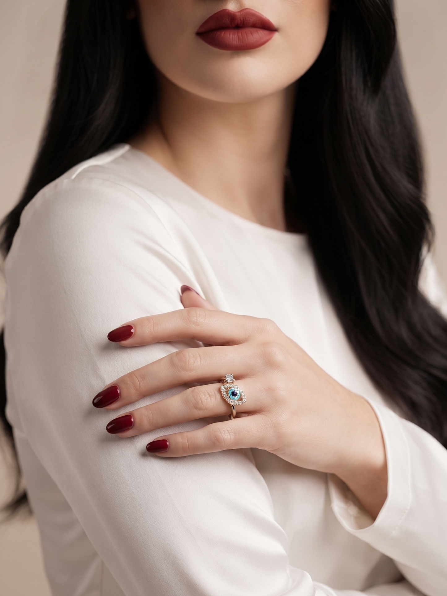 A close-up of a woman wearing a silver ring with a blue and white evil eye design, styled with a white long-sleeve top and dark red nail polish.