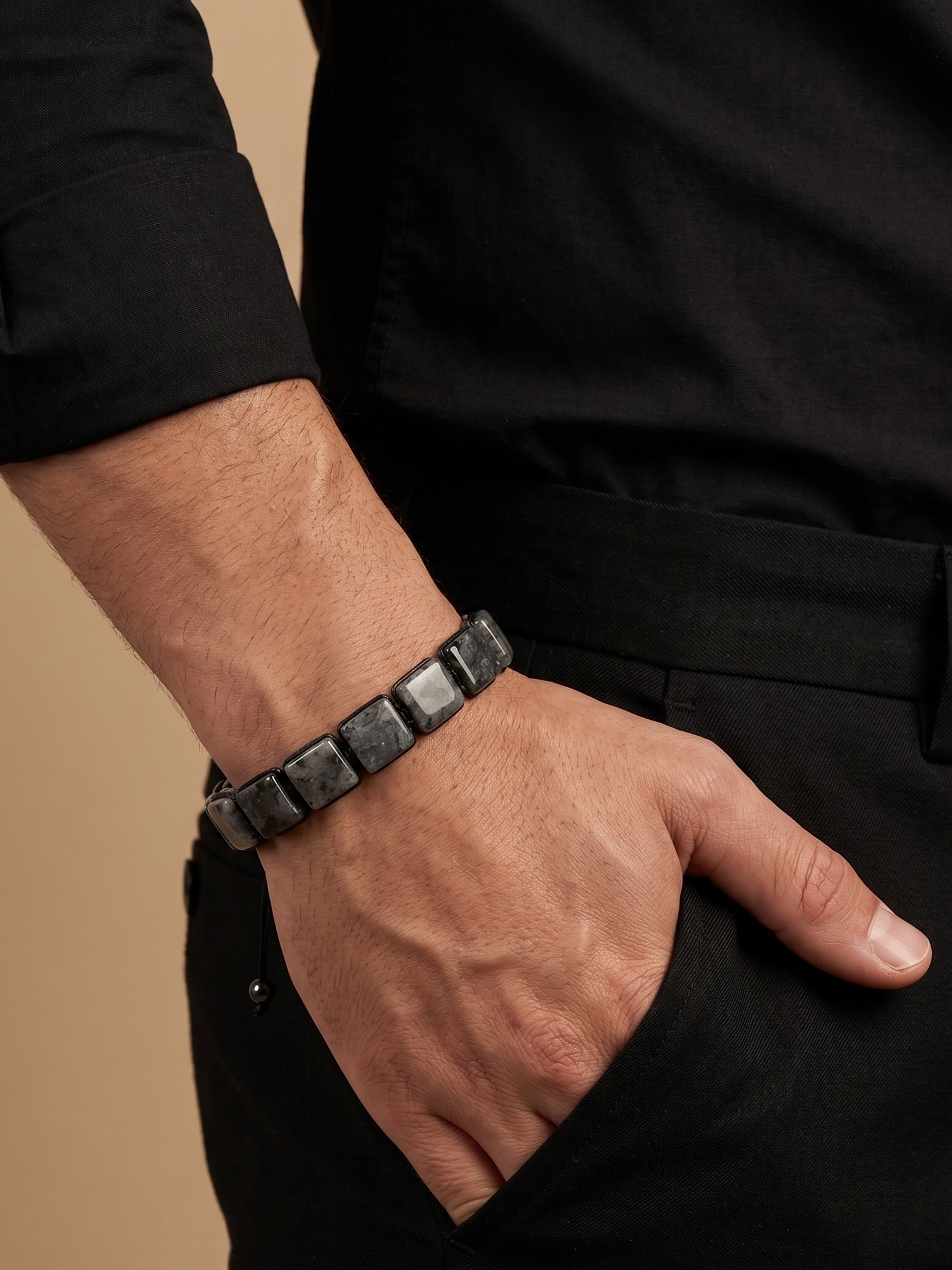 A close-up of a man's hand in his pocket wearing a dark gray stone bracelet, dressed in a black shirt and trousers against a warm beige background.